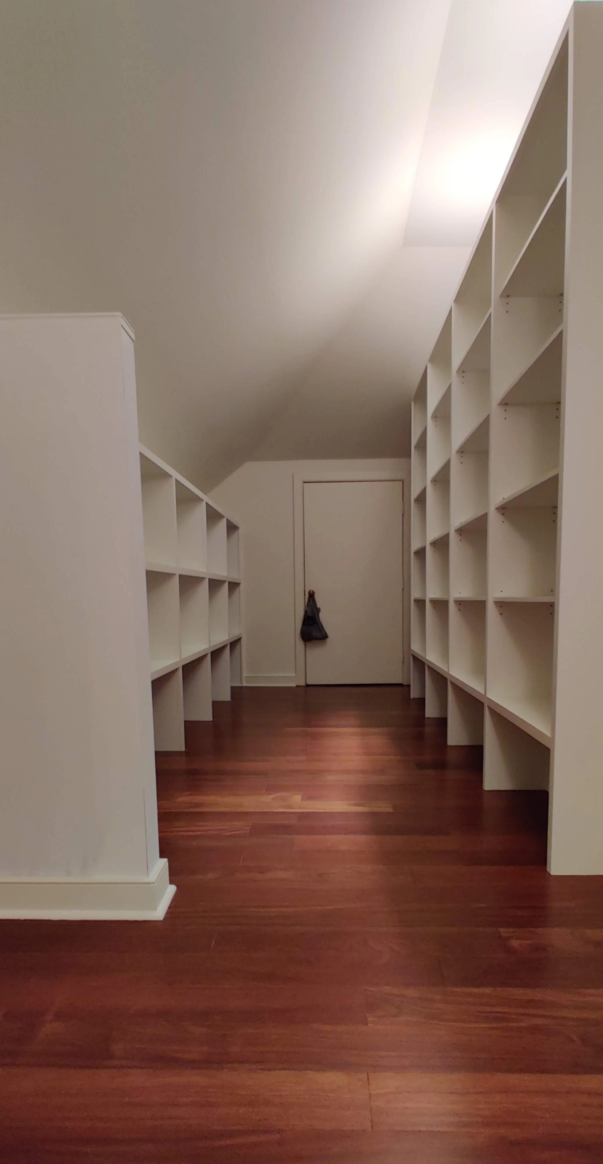 Empty walk-in closet with white built-in bookshelves on both sides and a wooden floor, door at the end with a black bag hanging on the door handle.