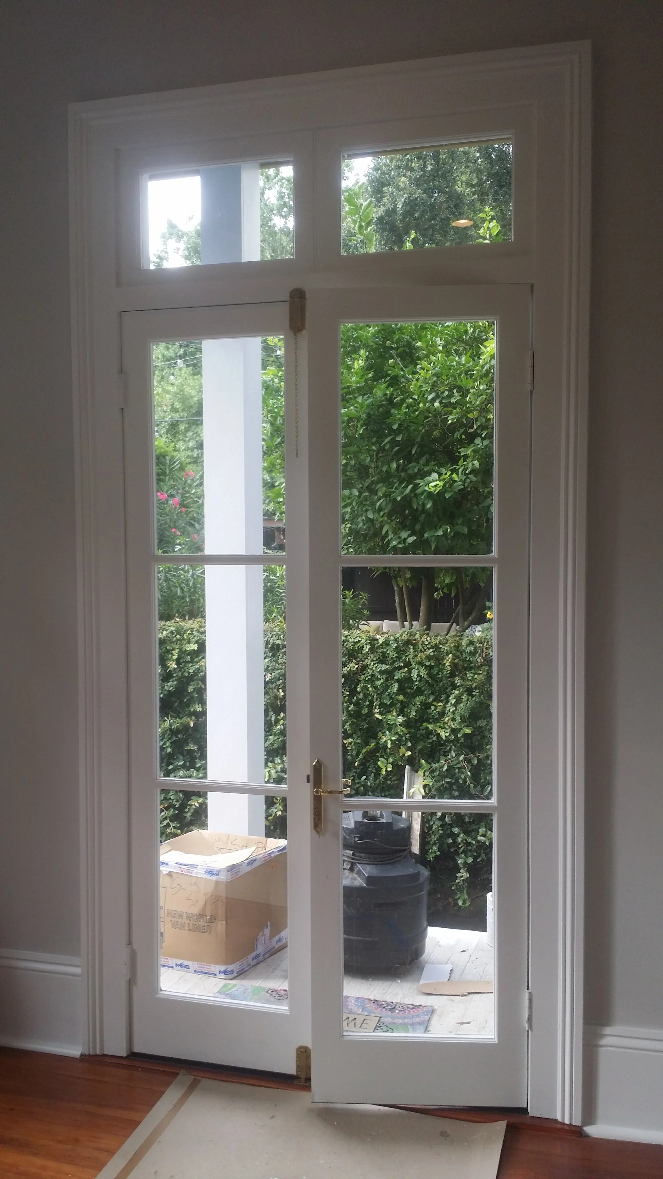 Interior view of a glass patio door leading outside, with window panes above and beside, showing outdoor greenery and a cardboard box on the porch.