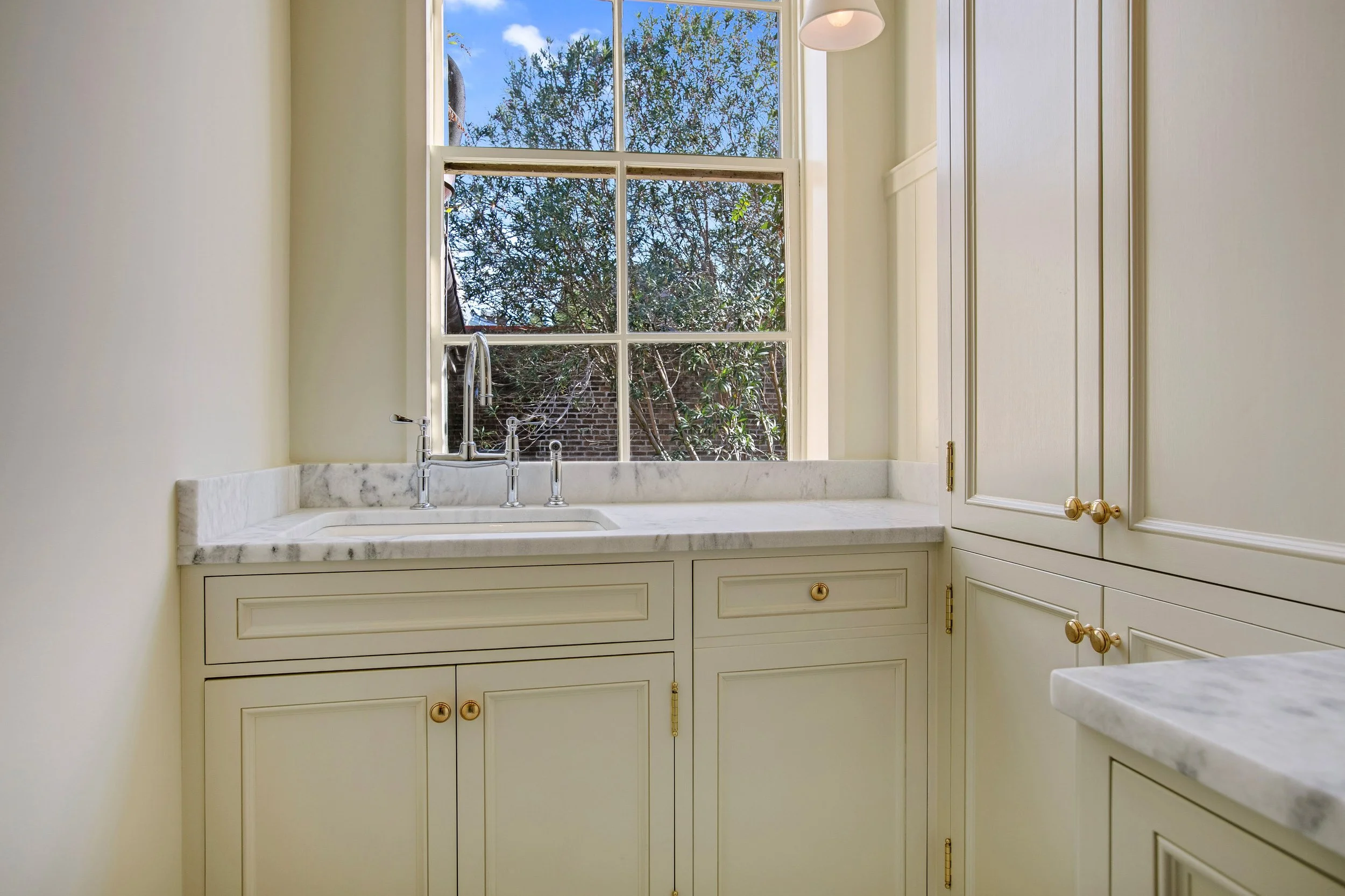 Kitchen with cream-colored cabinets, marble countertops, and a window overlooking trees and a brick wall, featuring a silver faucet.