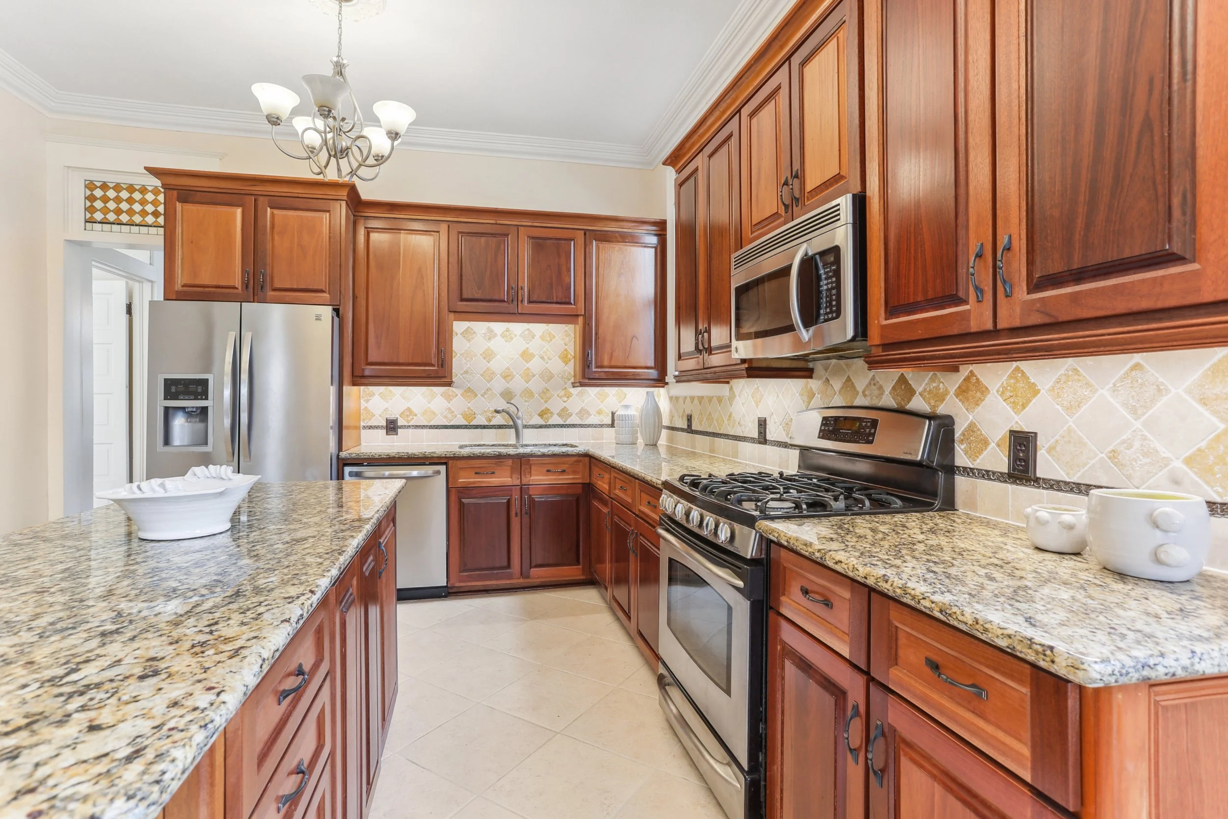 Kitchen with mahogany cabinetry, granite countertops, stainless steel appliances including a refrigerator, microwave, and oven, tiled backsplash, and a small chandelier.