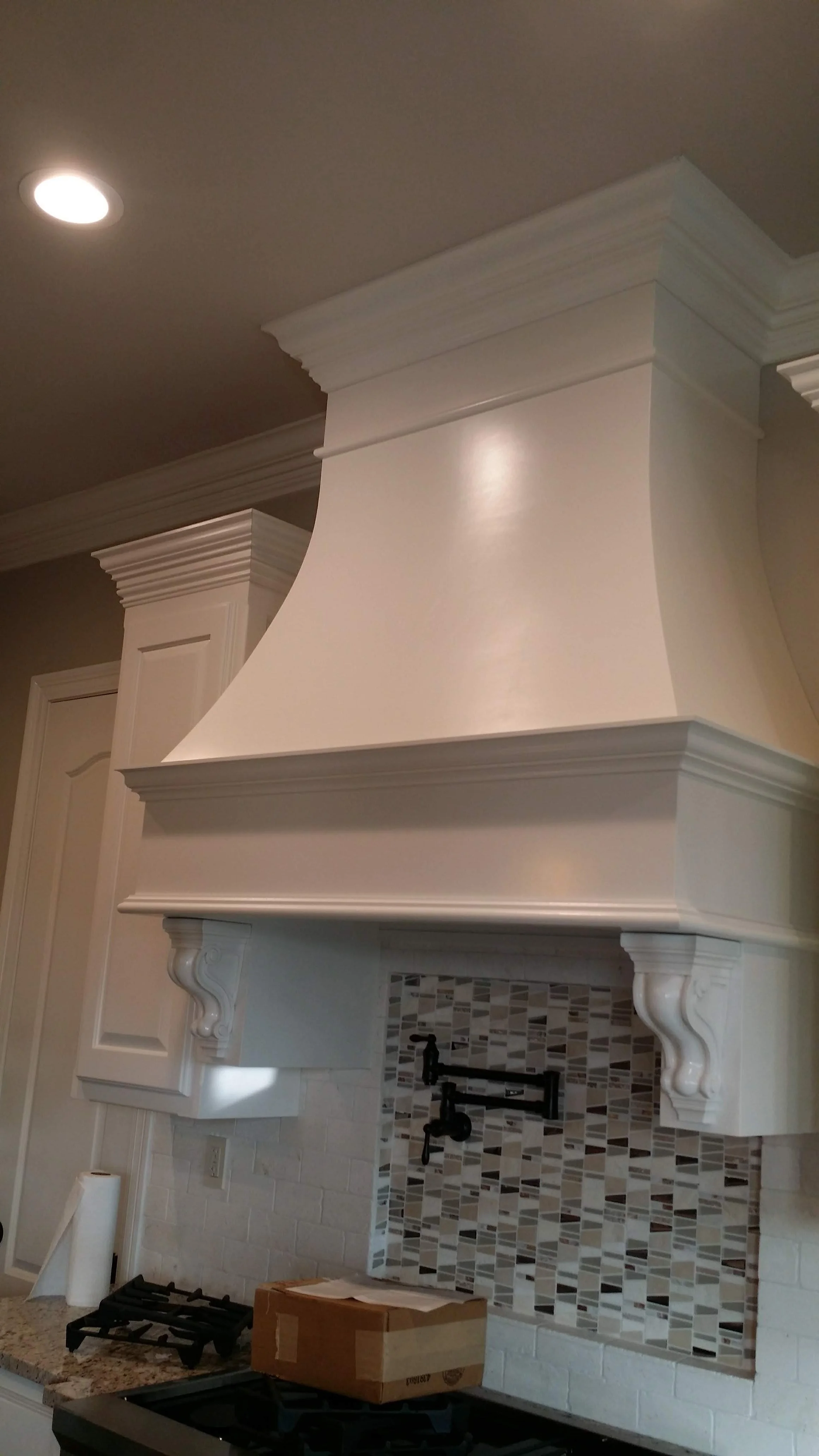 Close-up of a kitchen range hood with decorative molding and trim, above a stove area with a mosaic tile backsplash and black plumbing fixtures.