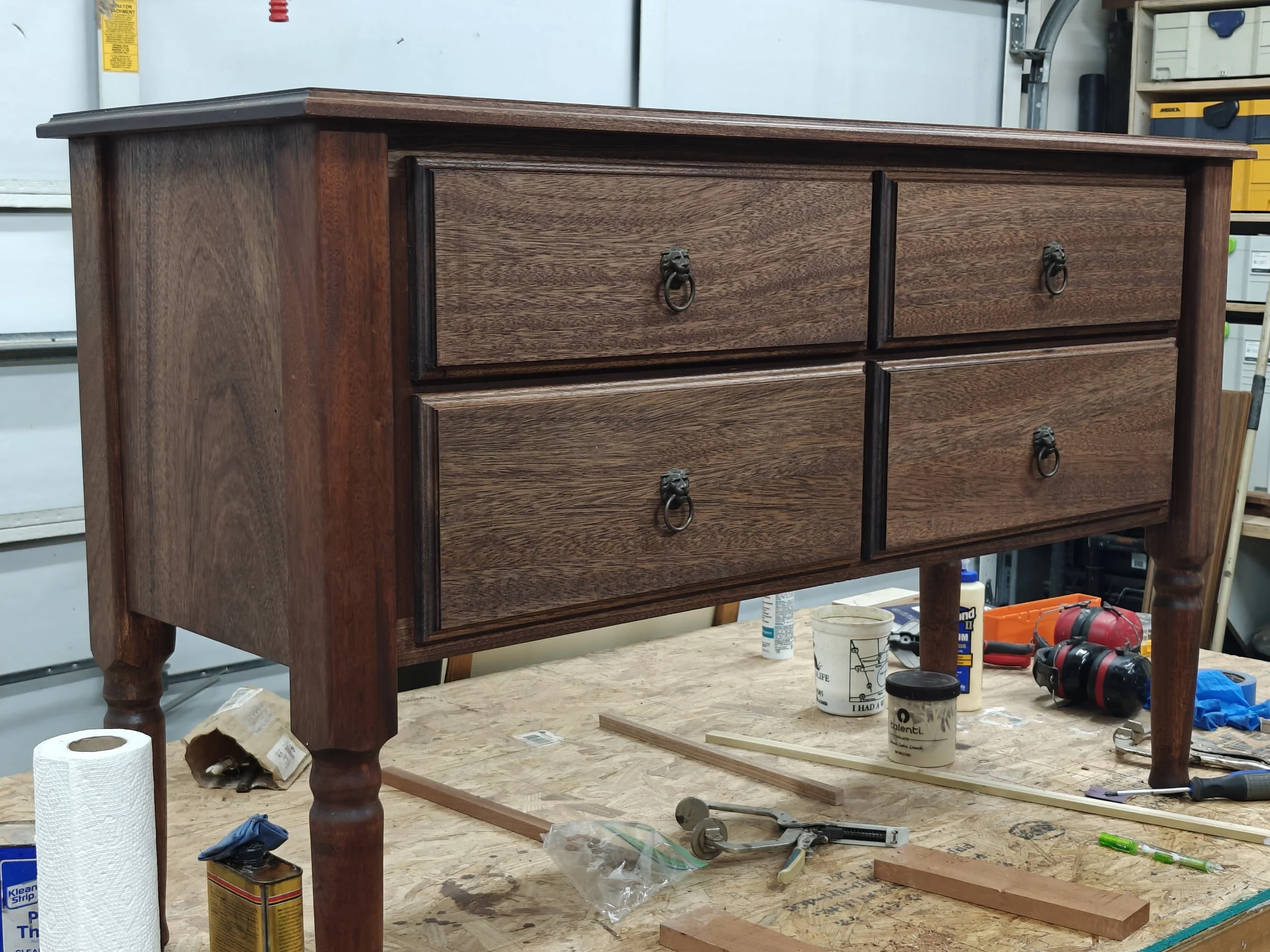 A mahogany dresser on a workbench surrounded by tools and supplies in a workshop.
