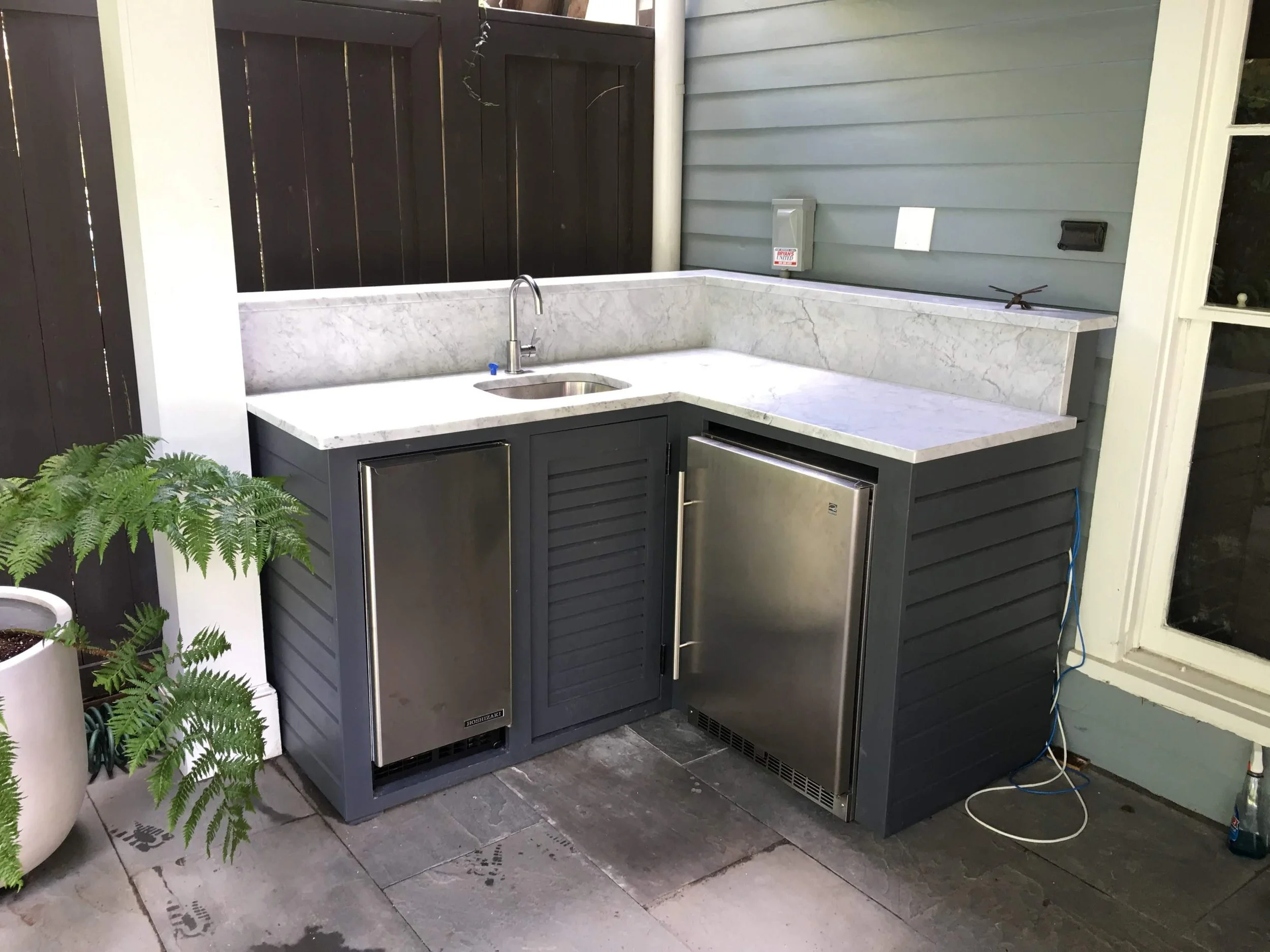 Small outdoor kitchen area with a gray cabinet with two stainless steel refrigerators, a marble countertop with a small sink, and a blue cord on the right side.