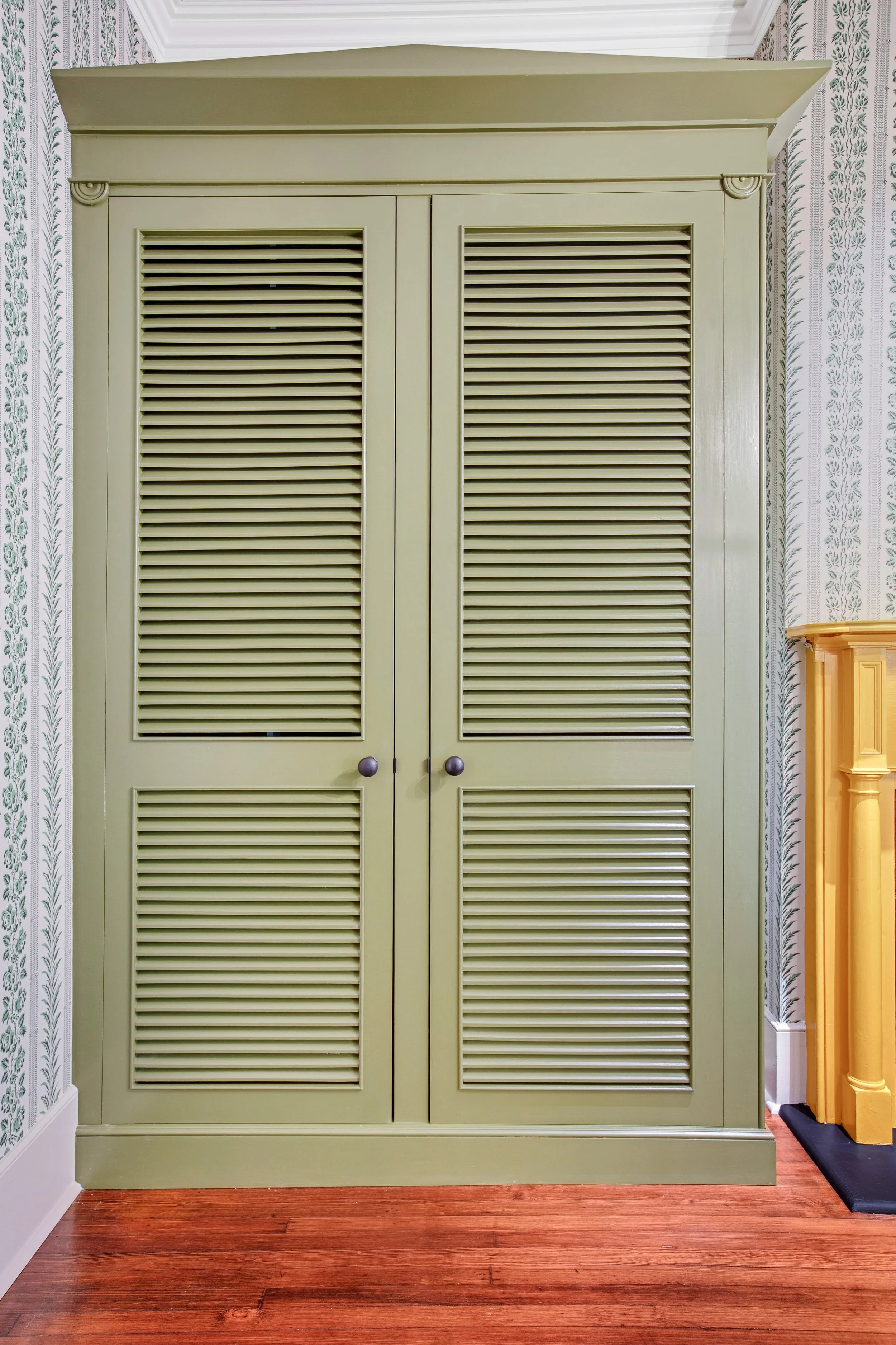 Custom built-in armoire with louvered doors with small round black knobs, set in a room with patterned wallpaper and wooden flooring.
