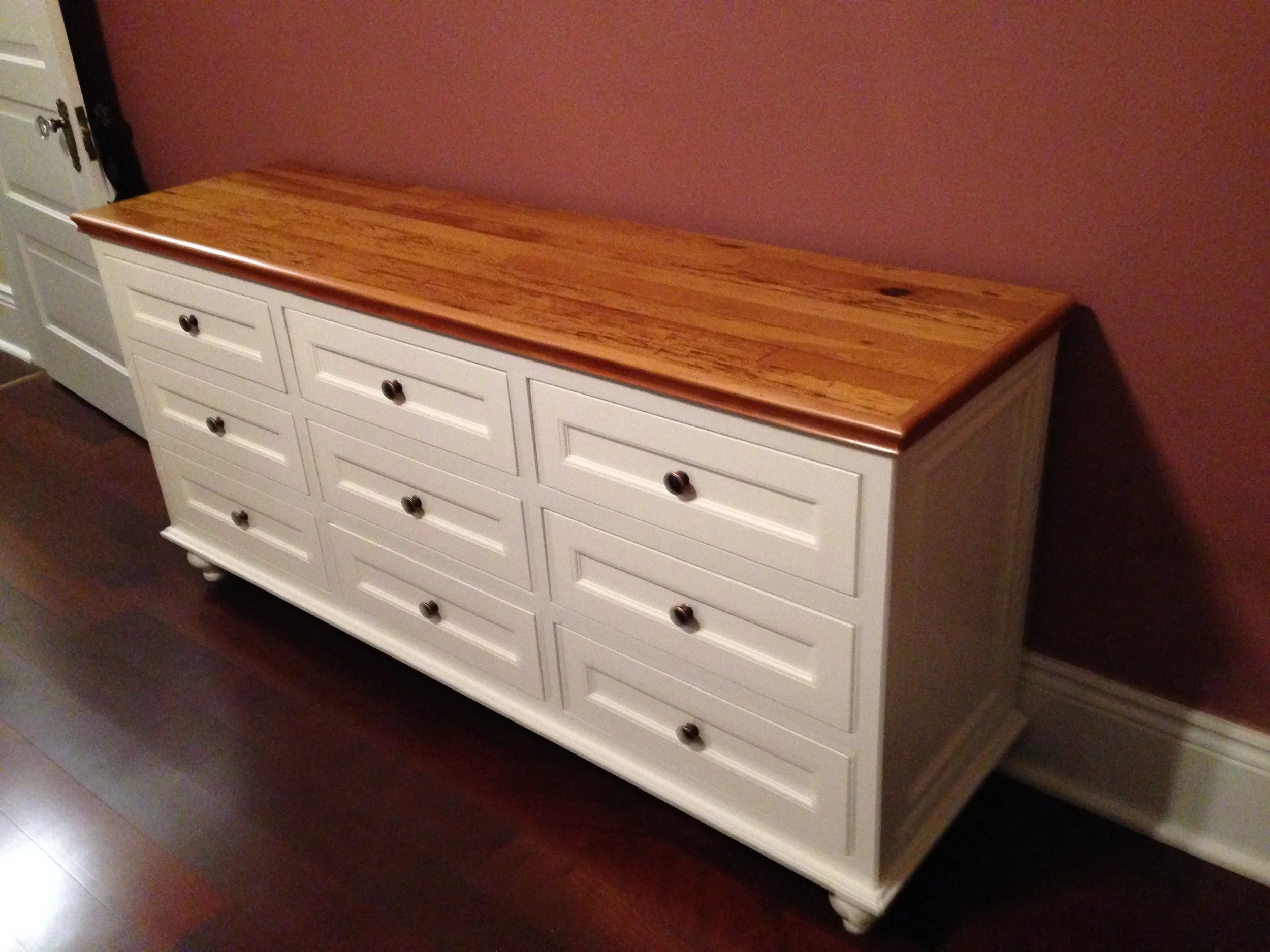 A white wooden dresser with nine drawers, nine dark round knobs, and a polished wooden top, placed against a pink wall on a dark hardwood floor.