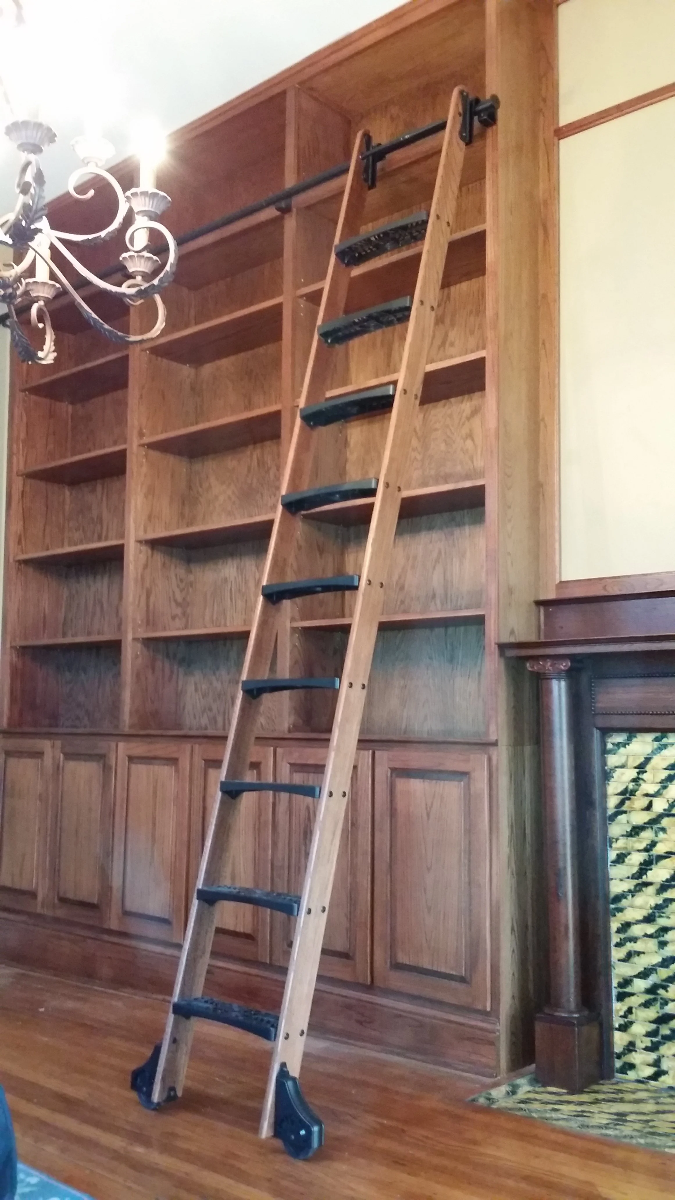 A rolling library ladder leaning against a large wooden bookshelf in a room with hardwood floors and a chandelier.