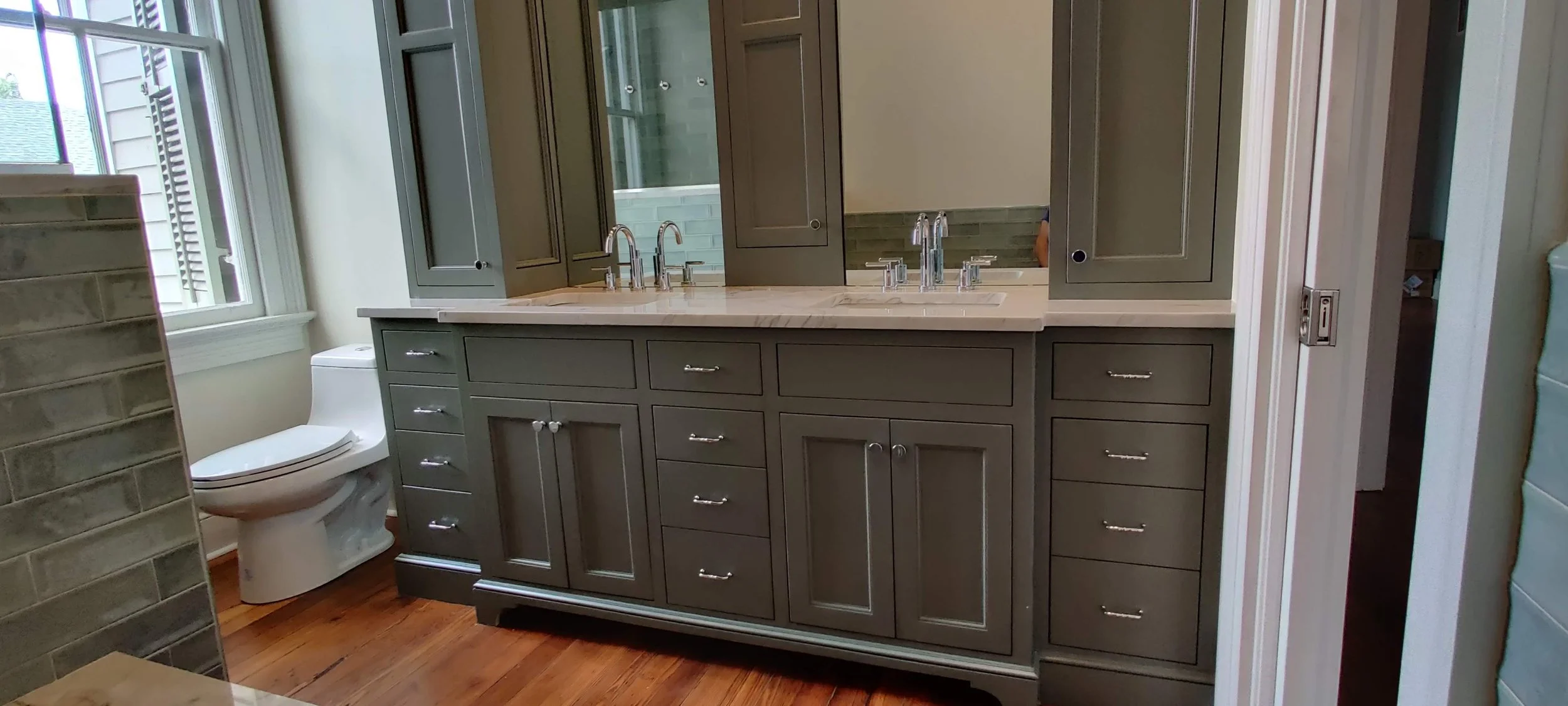A bathroom with a custom built gray vanity featuring multiple drawers and cabinets, a marble countertop with two sinks and shiny silver faucets, a large mirror above the vanity, a window on the left side, a toilet near the window, and wood flooring.