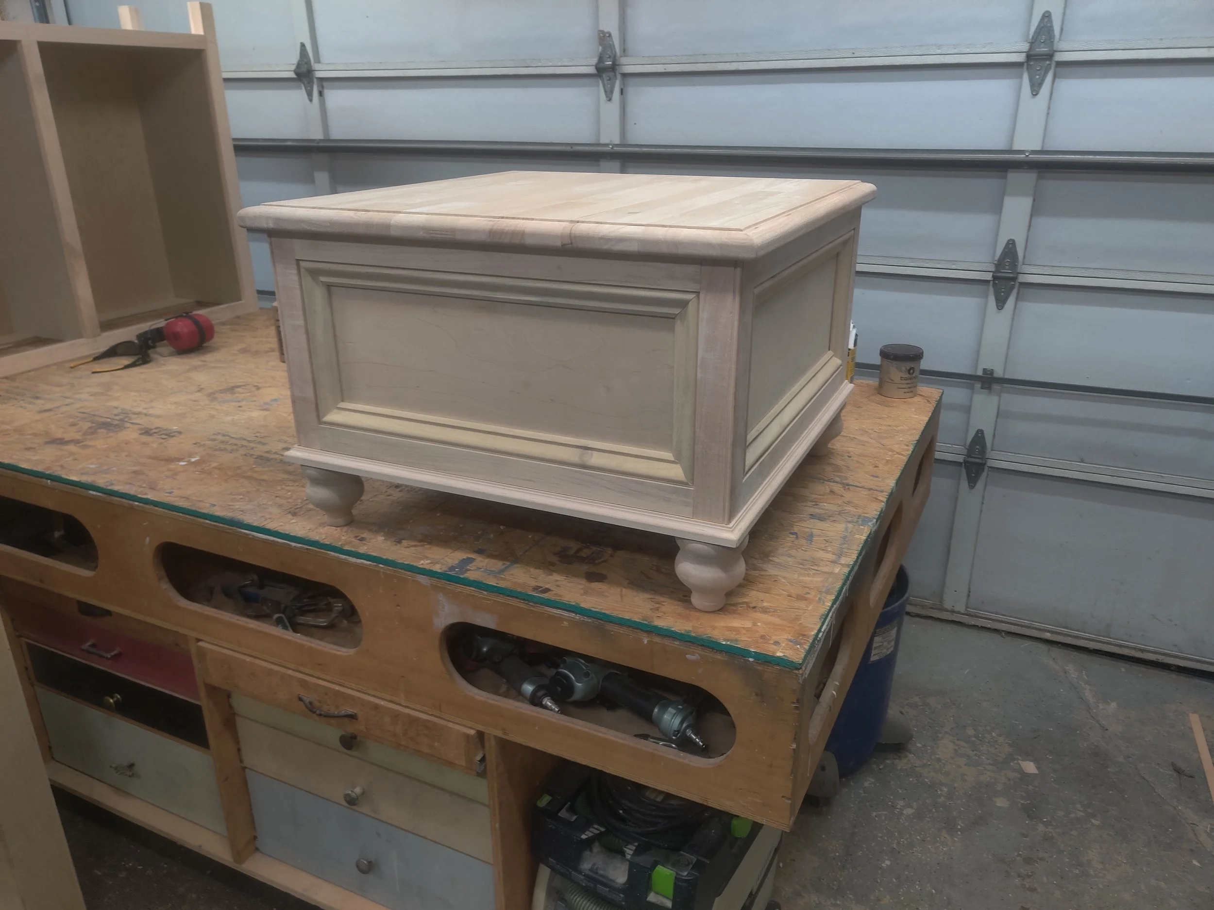 A wooden chest with a light finish and decorative molding, sitting on a workbench in a woodworking shop.
