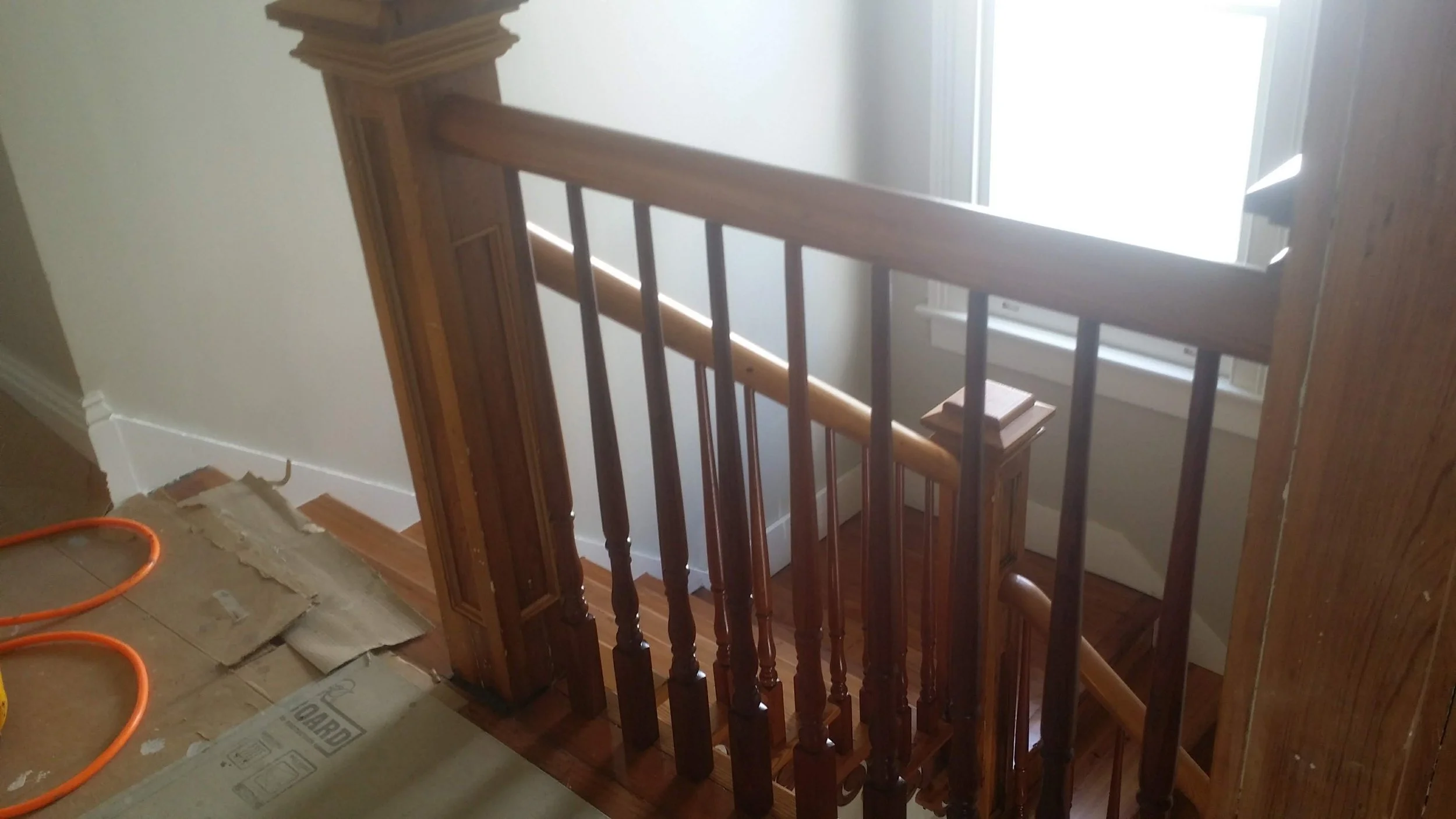 Wooden staircase with handrail and balusters next to a window, with some tools and cardboard on the floor.