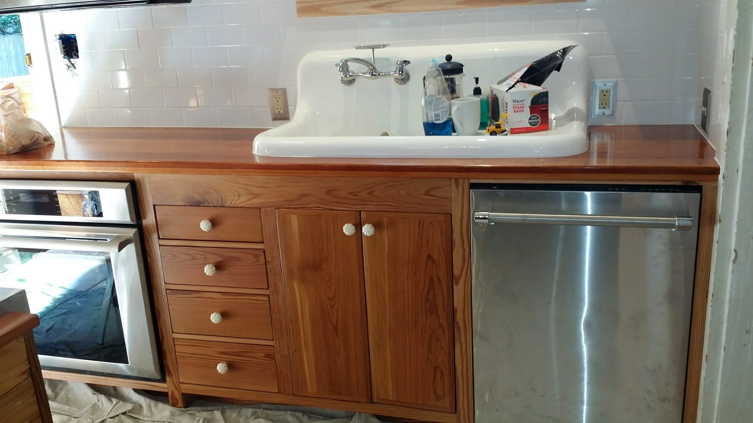 Kitchen with reclaimed heart pine cabinets, a white farmhouse sink with items on the counter, a stainless steel dishwasher, and a white tiled backsplash.