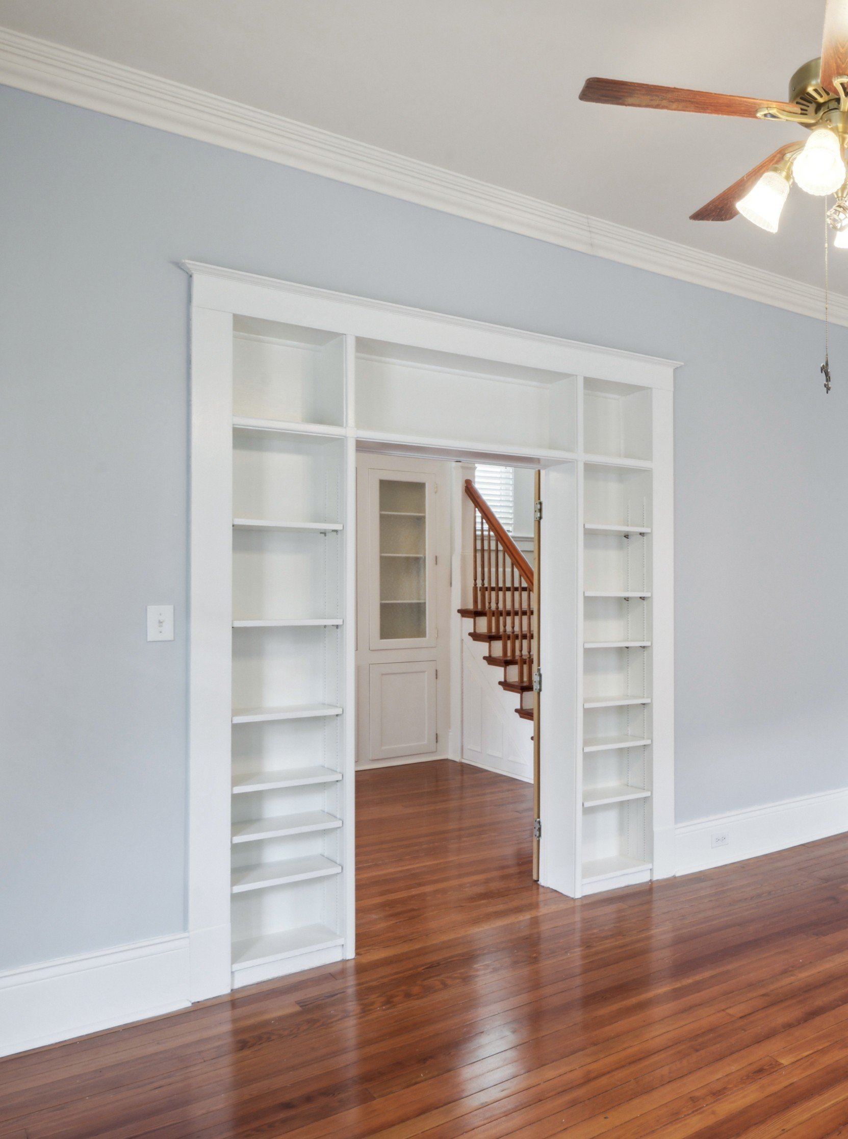 Interior view of a room with light blue painted walls, white trim, and hardwood floors. It features a white built-in bookshelf with open shelves surrounding an open doorway. Through the doorway, a staircase with wooden steps and handrail is visible, 