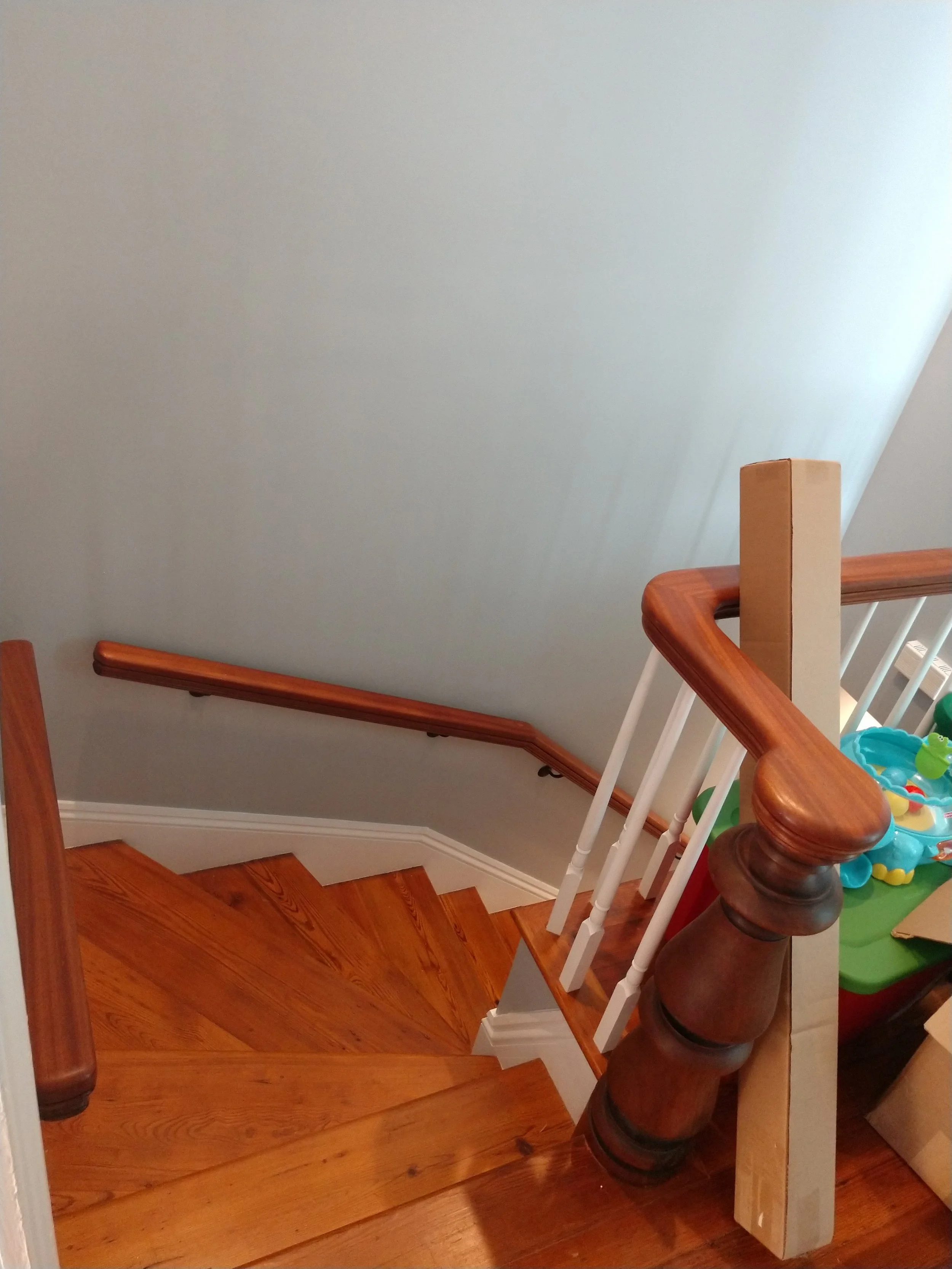 Wooden staircase with a curved handrail and white spindles leading to the lower floor, with a wall on the left and a newel post on the right. Part of a cluttered space with toys and boxes.