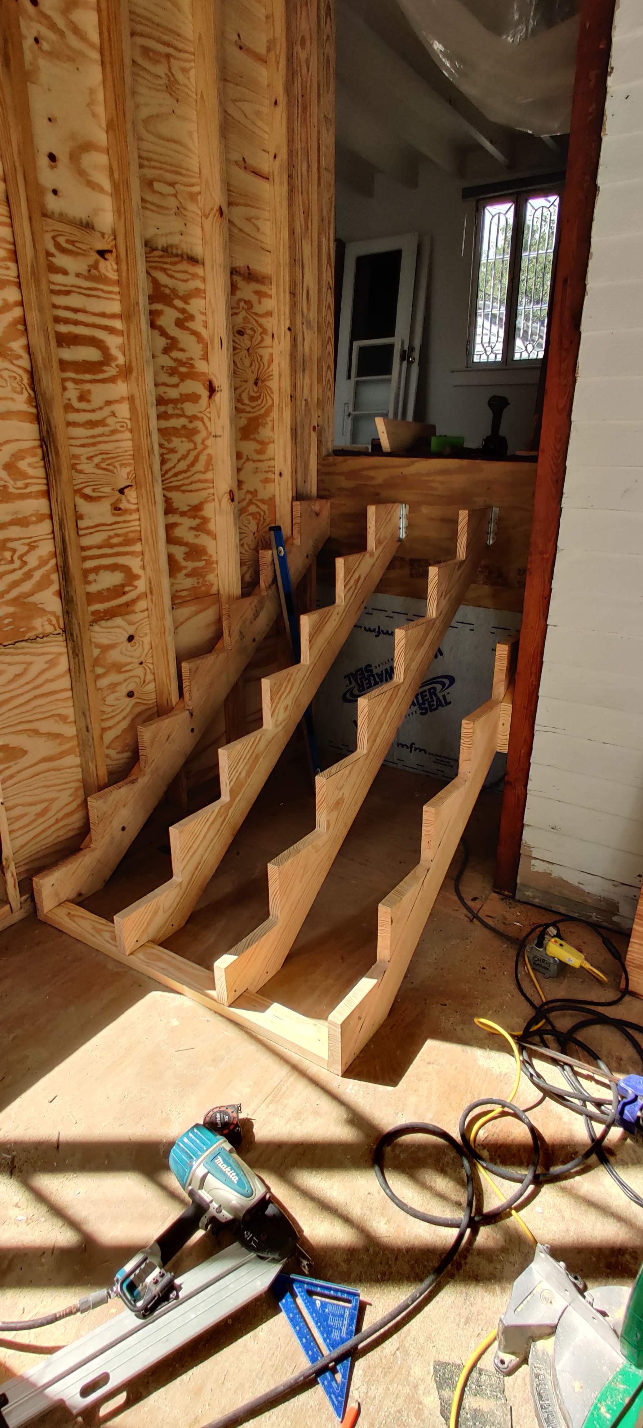 Wooden staircase construction in progress inside a building. Tools and construction materials are visible around, with part of the interior wall and window in the background.