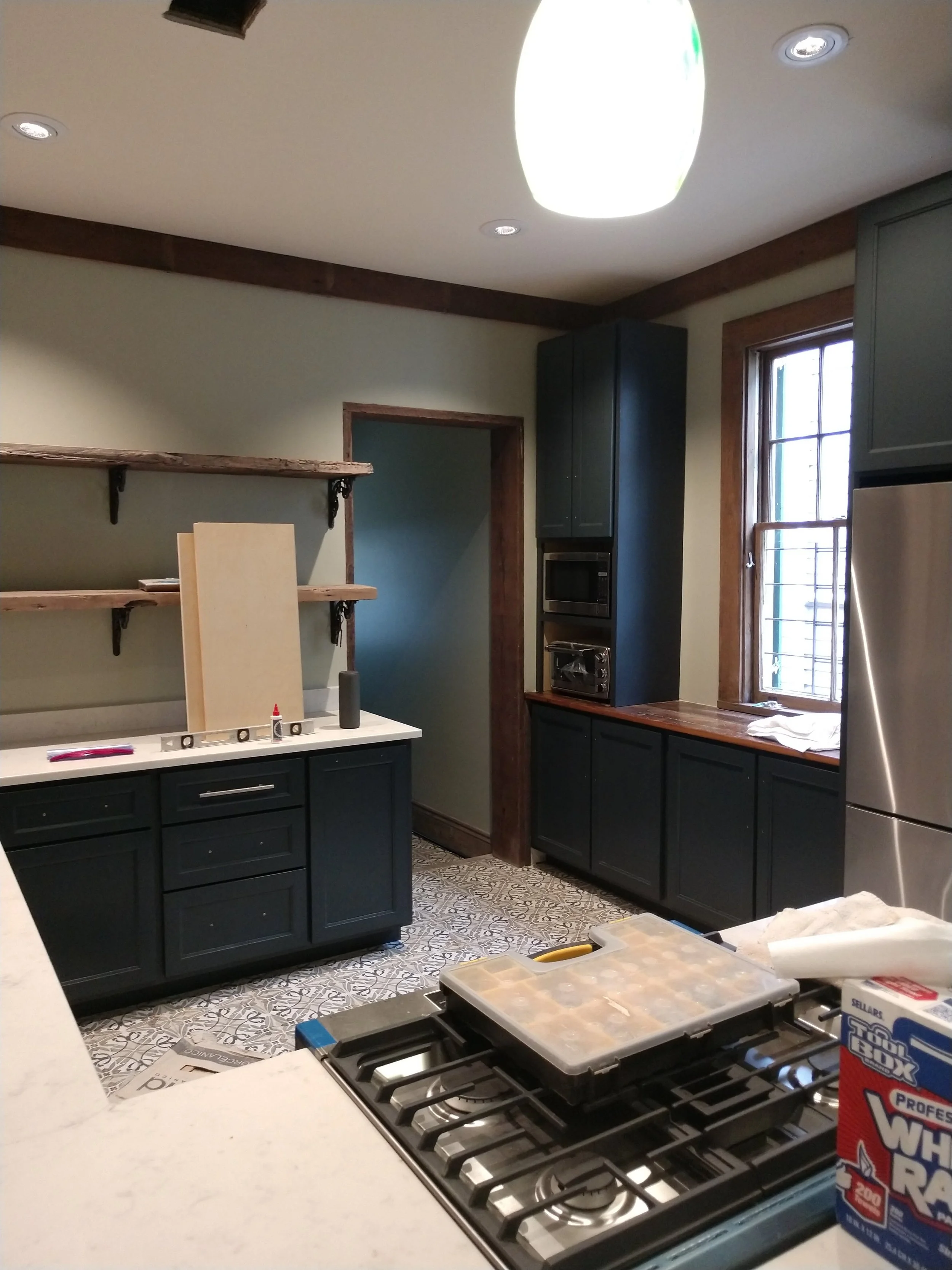 Kitchen under renovation with black cabinets, patterned floor tiles, and open shelves. A window with a wooden frame, a microwave, and toaster built into tall cabinets are visible. There are supplies and tools on the counter, including a box of trash 