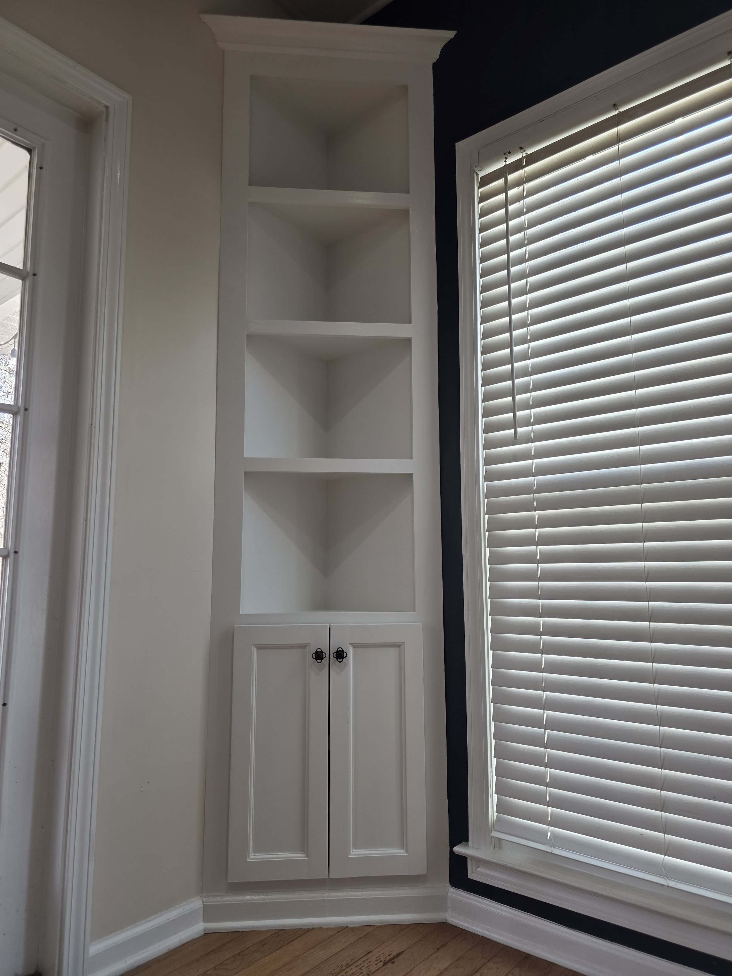 Built-in white corner cabinet with open shelves and closed doors next to a window with white blinds indoors.