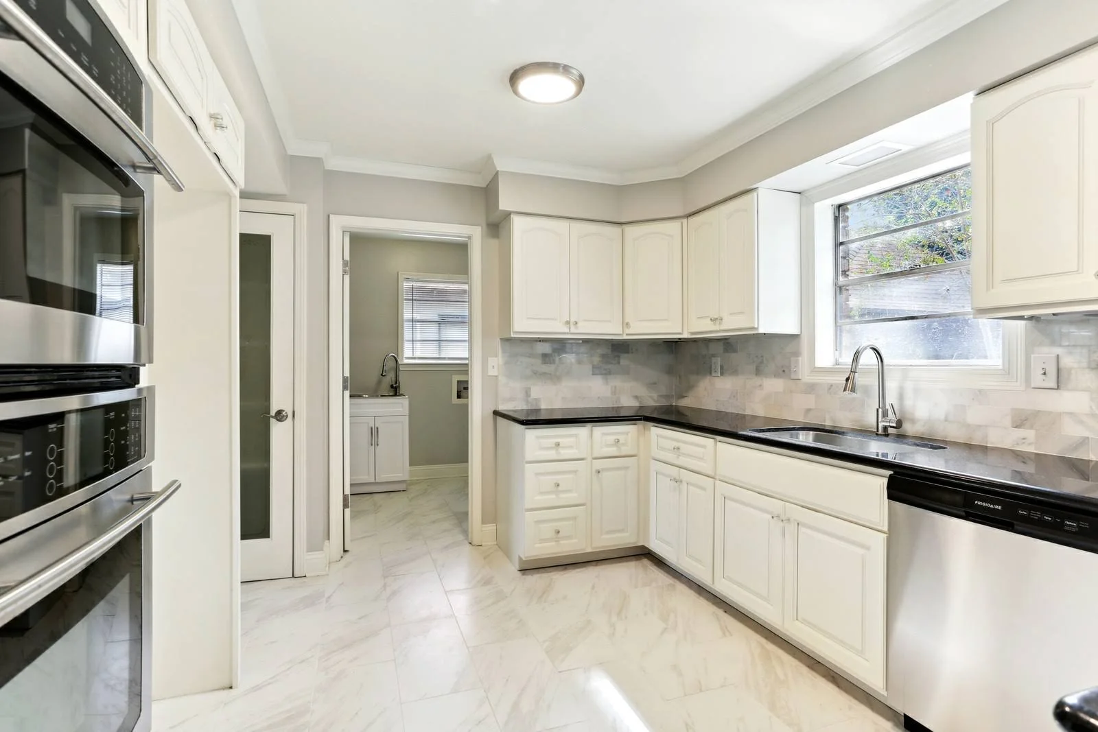 custom Kitchen with white cabinets, black countertops, marble floor and backsplash, stainless steel appliances, and a window above the sink.