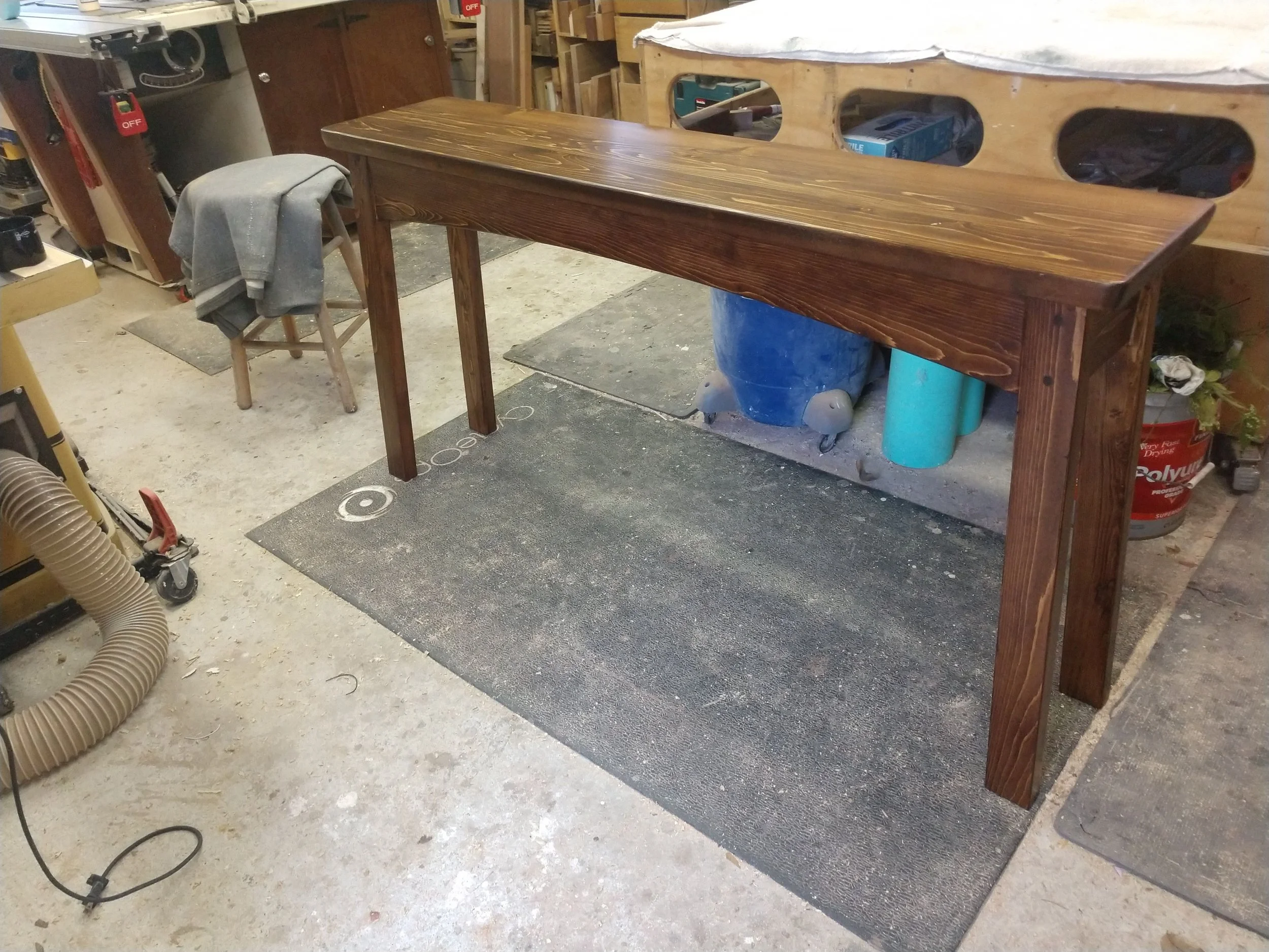A woodworking workshop with a new wooden table in the foreground. The table is dark stained with clean lines. A gray cloth is draped over a small wooden stool beside the table. Various tools and equipment are visible around the workshop, including a 