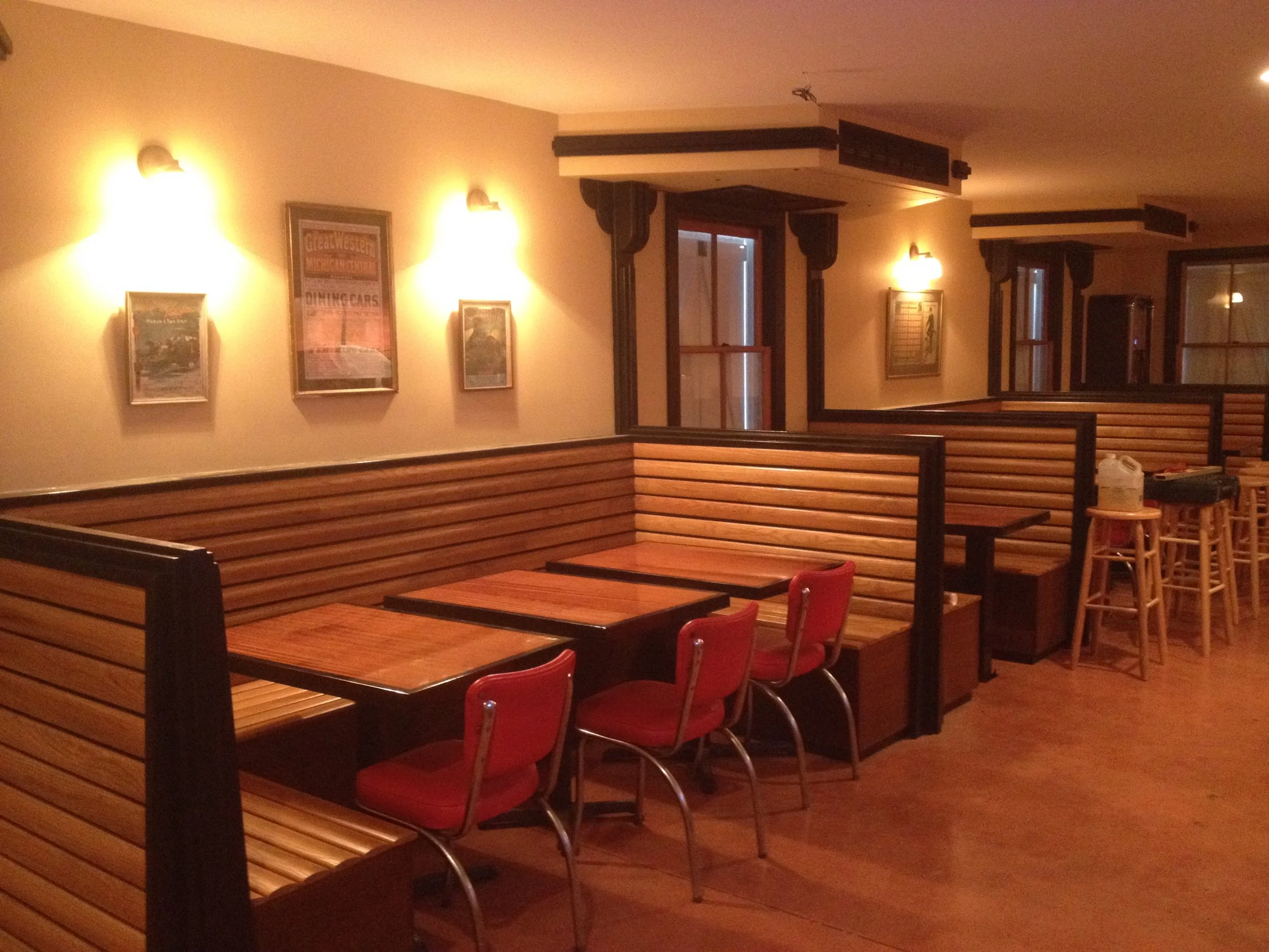 Empty restaurant booth with wooden benches and red chairs, framed posters on beige walls, warm lighting, and wooden flooring.