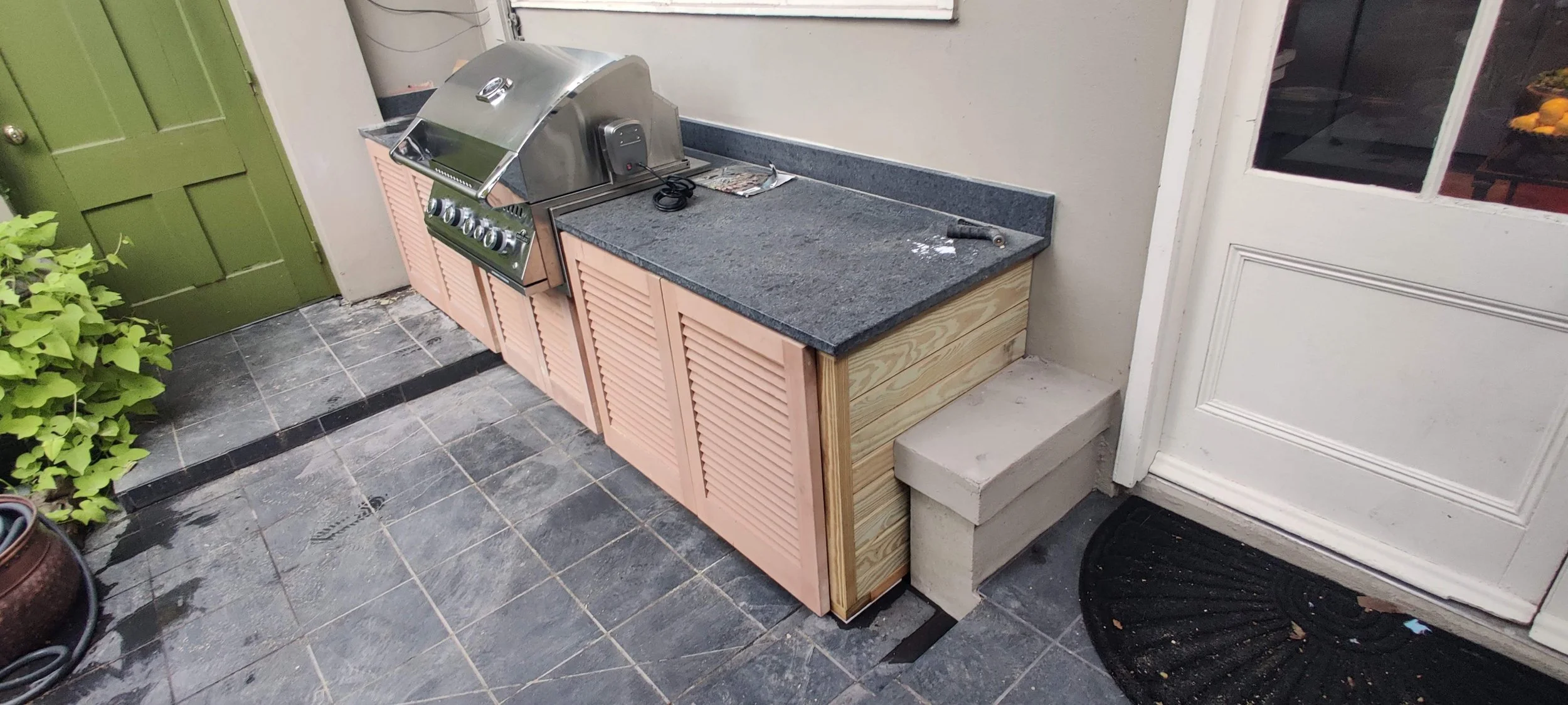 An outdoor kitchen area with a black countertop, pink cabinets, a stainless steel grill, and some tools on the counter. 