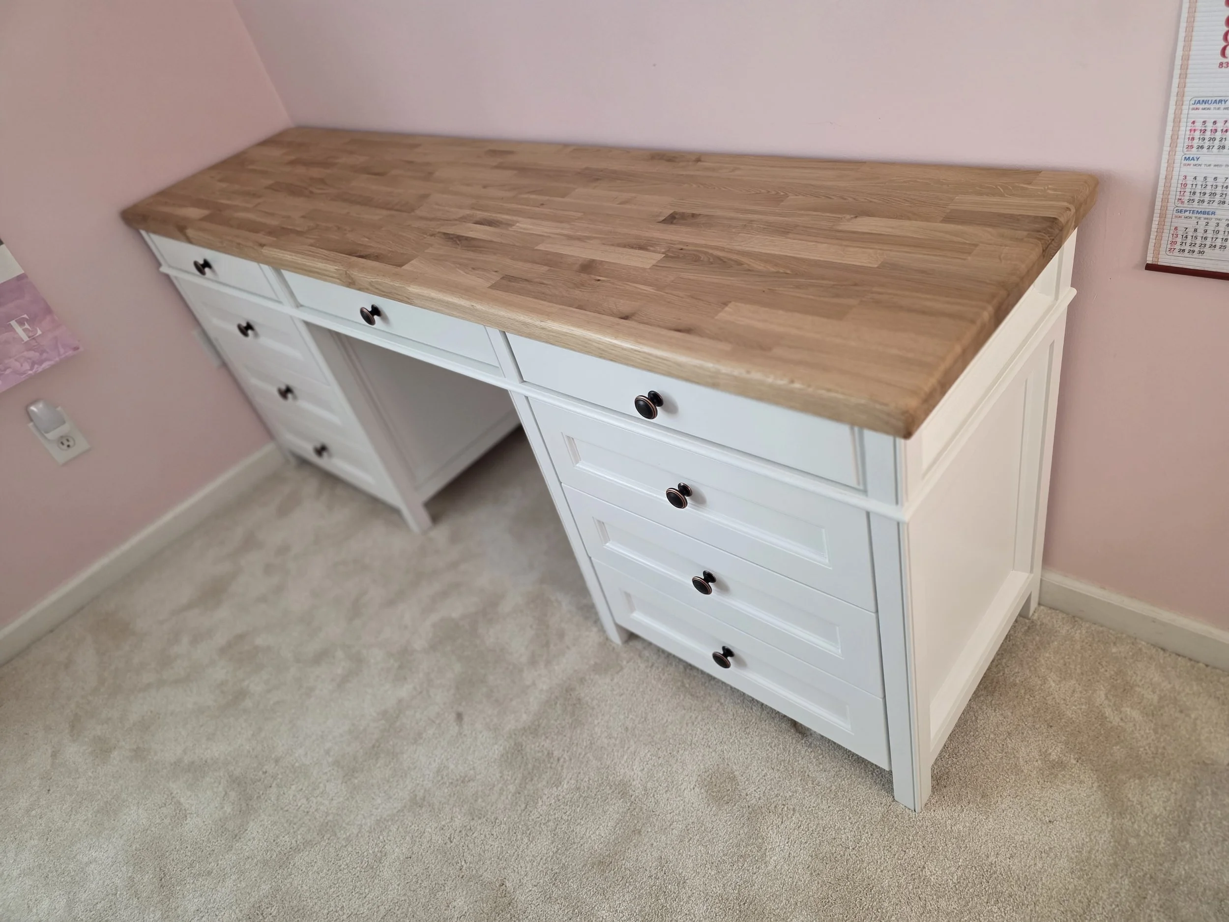 White wooden dresser with multiple drawers and black knobs, with a wooden tabletop, positioned against pink walls in a room with beige carpet.