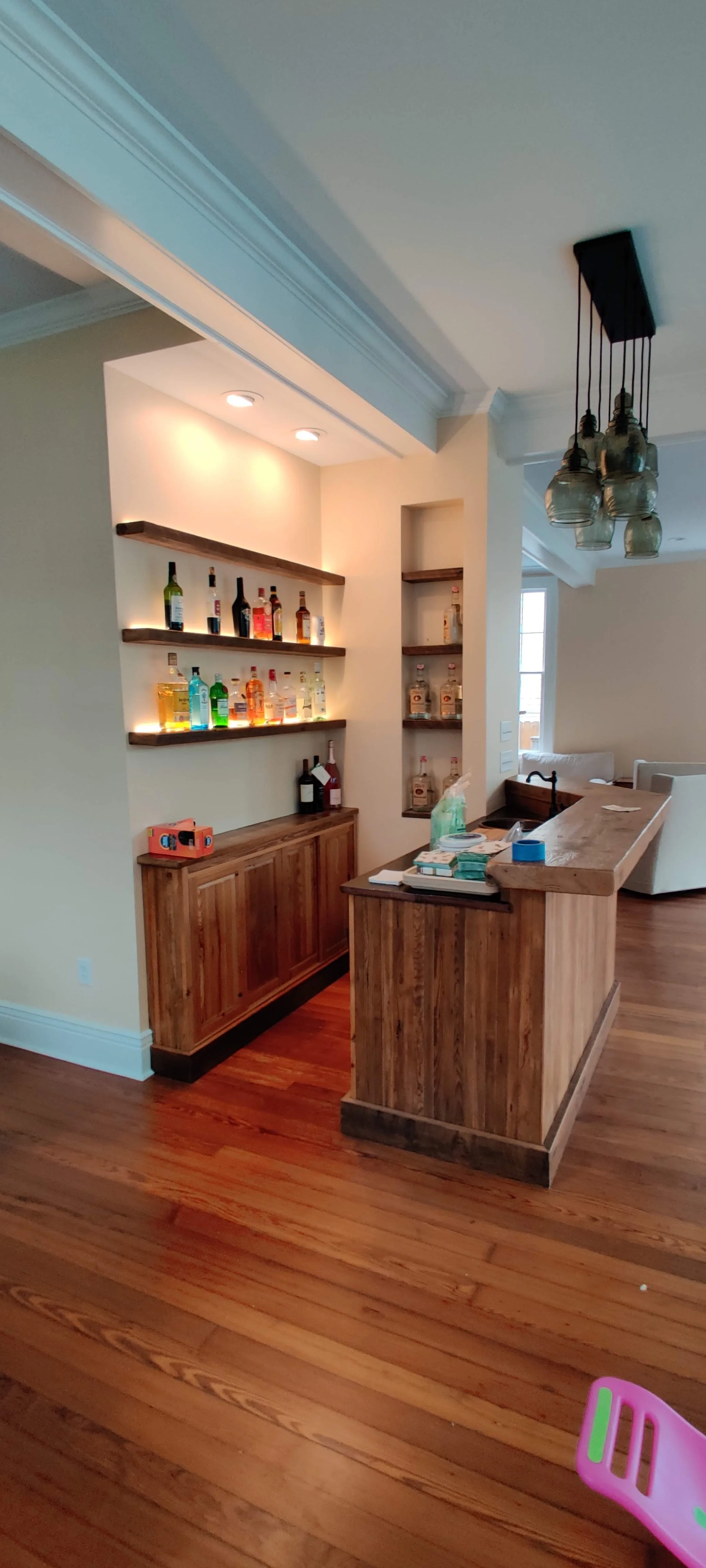 A built-n wet bar with wooden shelves holding various liquor bottles, under recessed lighting, with a wooden counter and a wooden cabinet below. There are some items on the counter, and a modern chandelier with glass jars hanging above. Part of the l