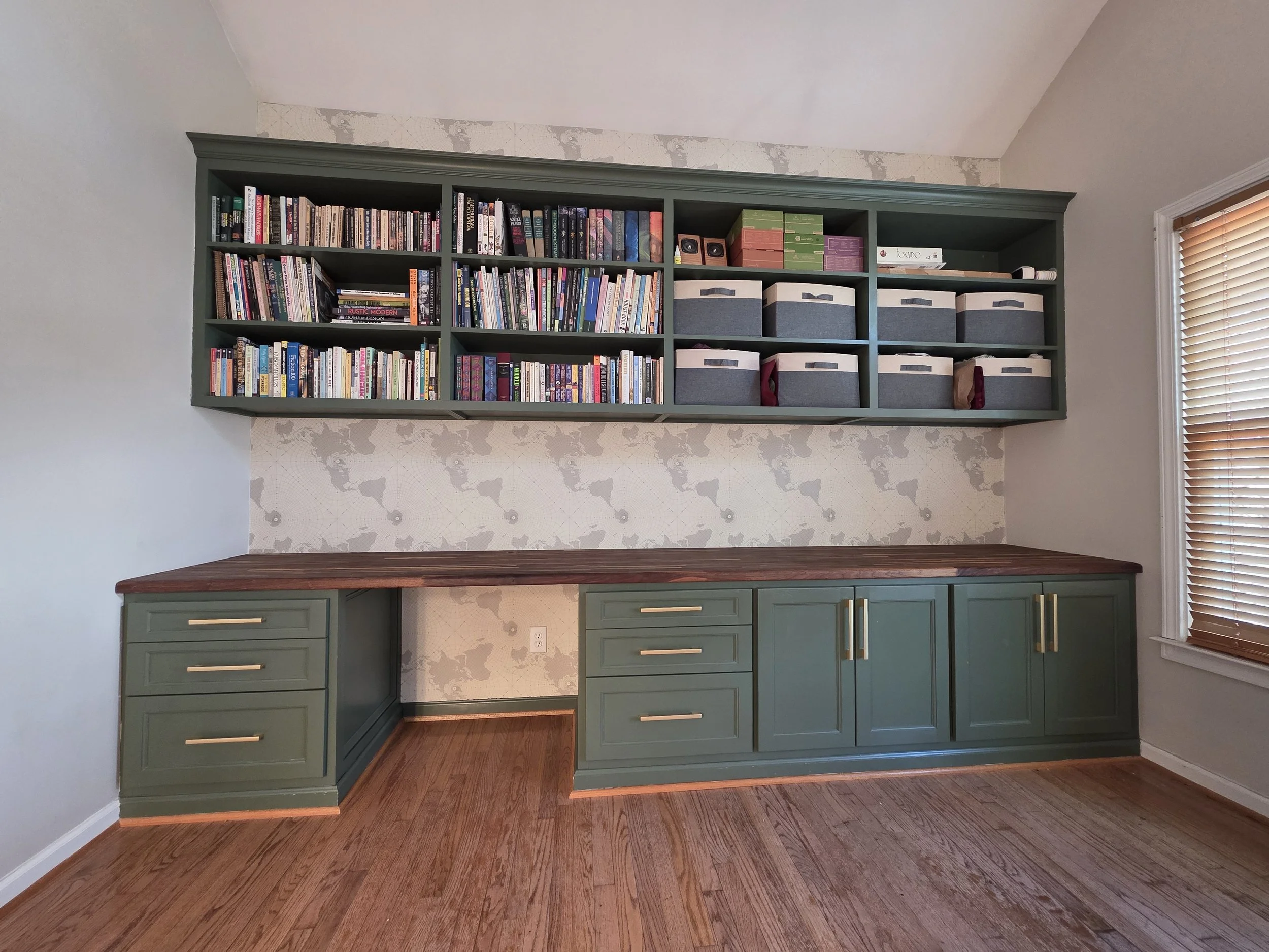 Built-in desk with green cabinetry, a wooden desk, a built-in bookcase filled with books, storage boxes, and a window with wooden blinds.