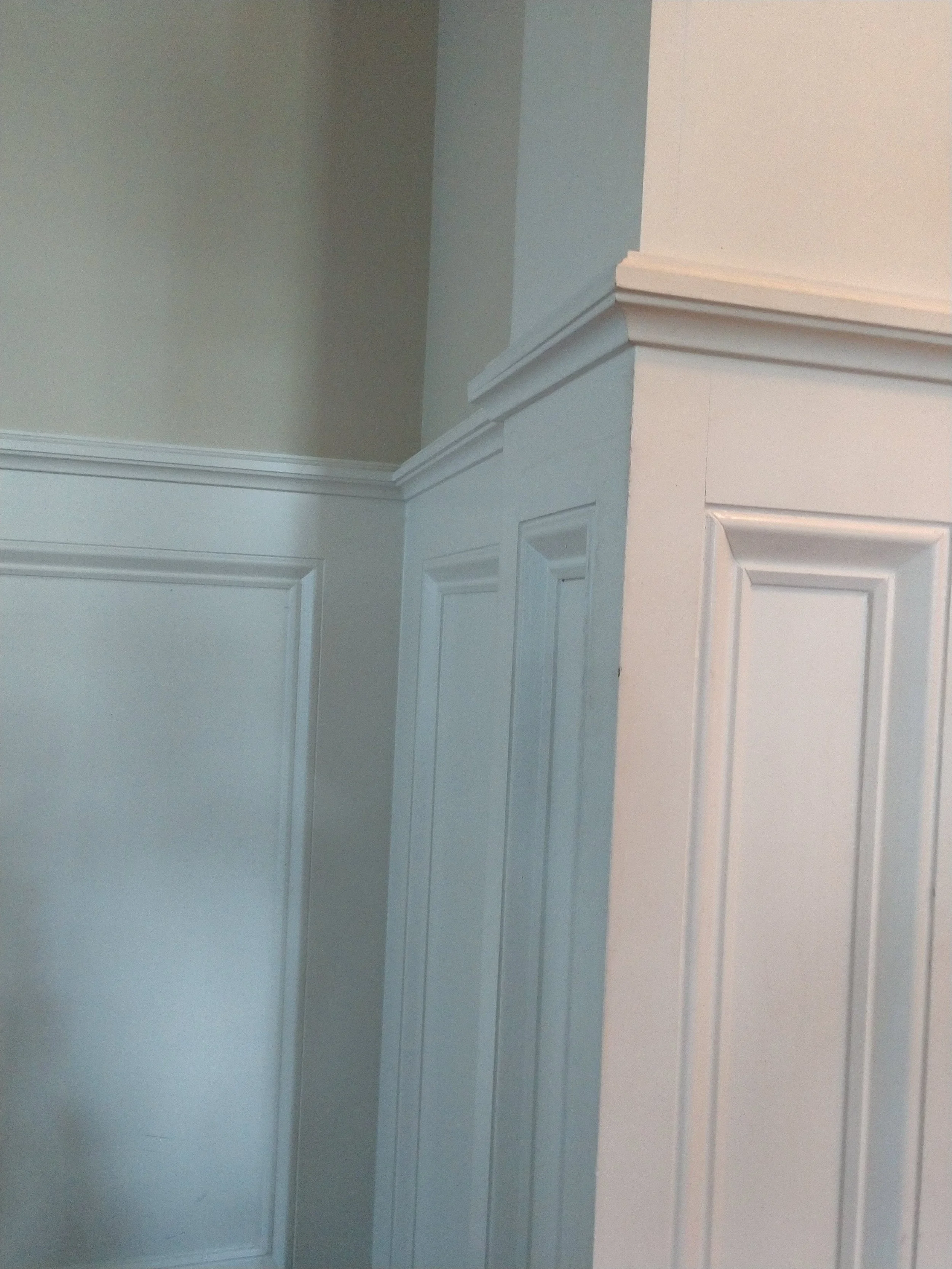 Corner of a room with white wainscoting and beige upper wall, featuring decorative molding and cornices.
