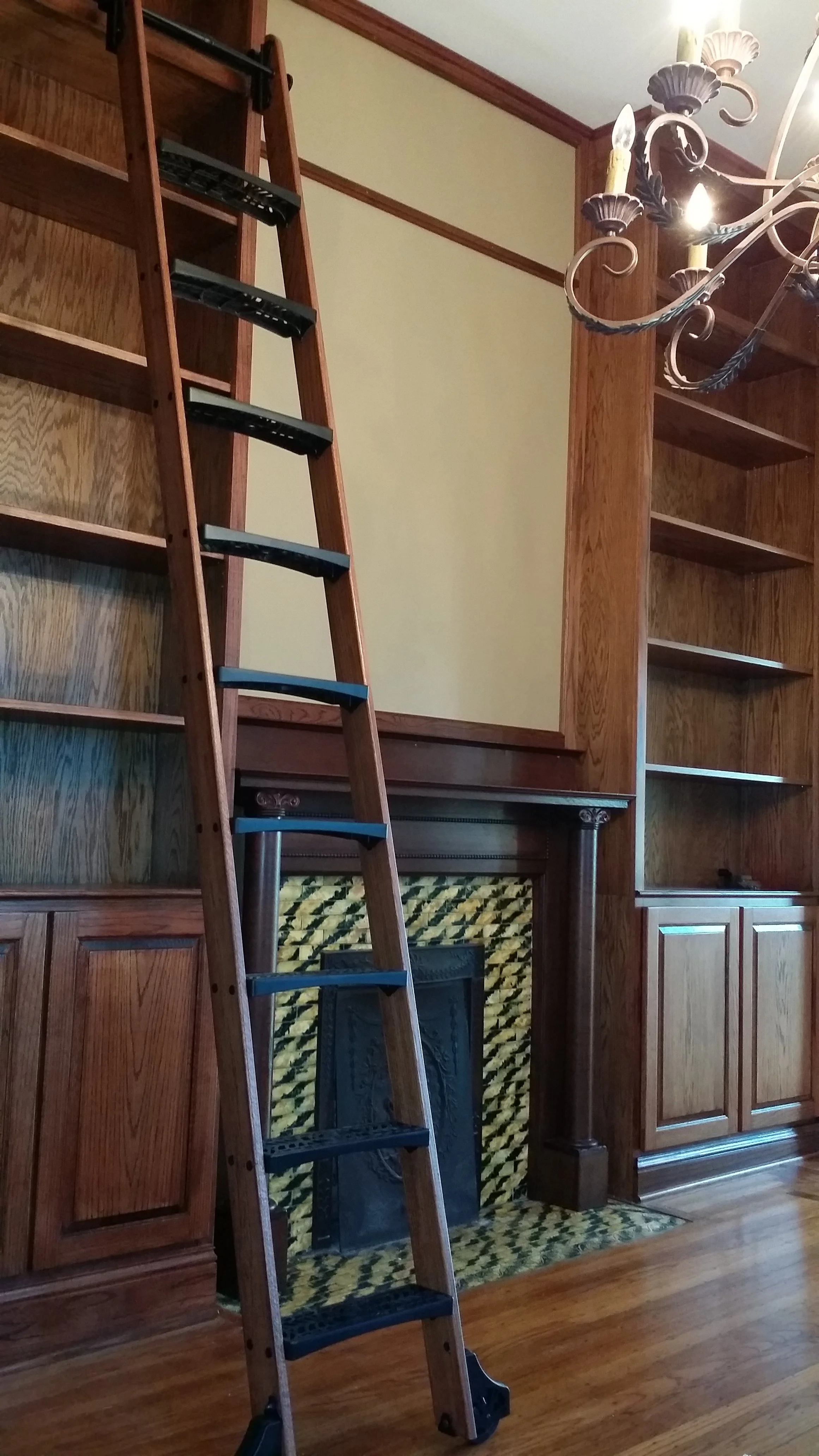 Wooden ladder leaning against built-in wooden library and a fireplace with a decorative tile surround, in a room with hardwood floors and a chandelier.