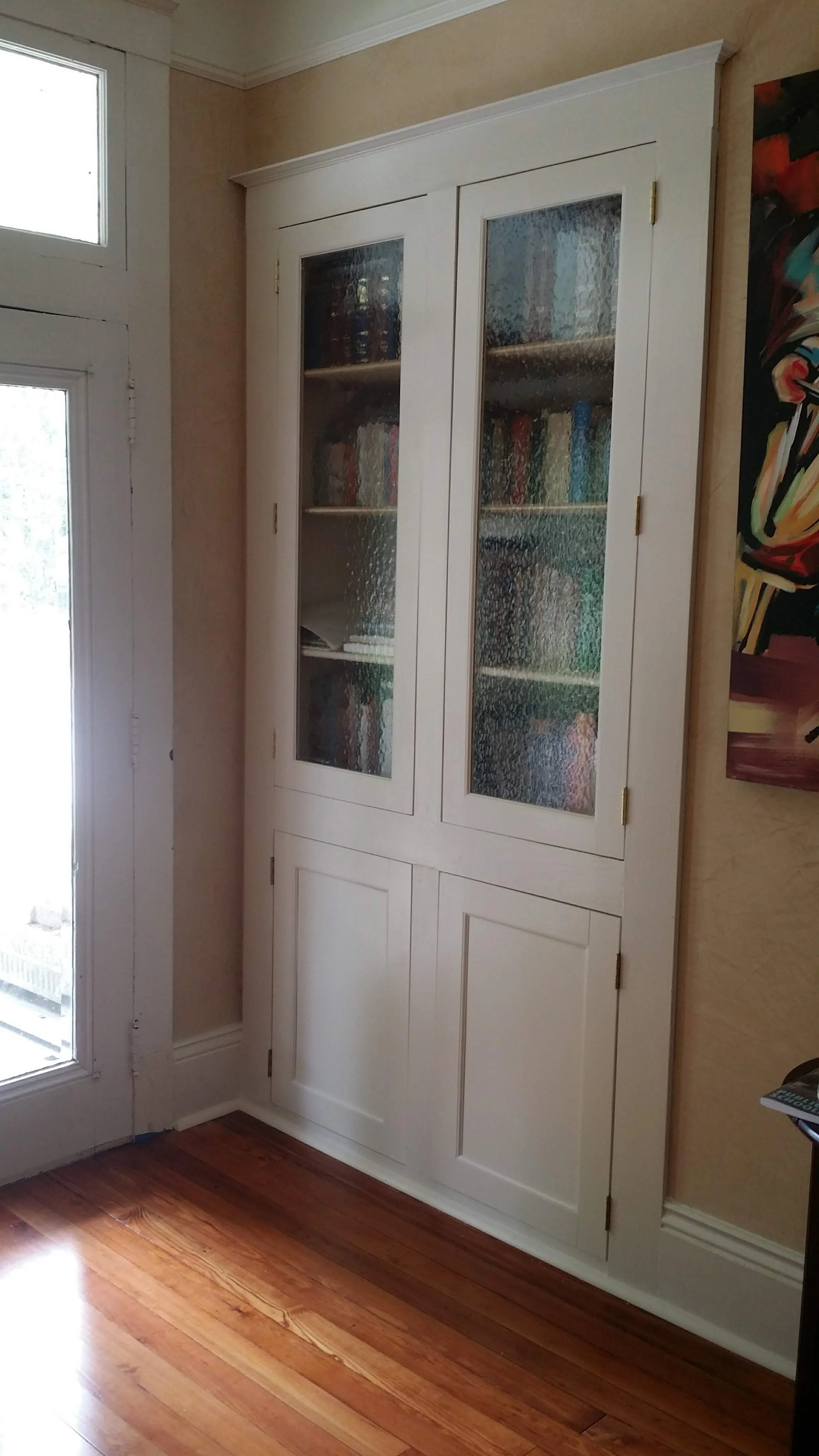 A white built-in cabinet with glass-paned doors containing books, situated on a beige wall near a glass door with sunlight coming in, and a wooden floor.