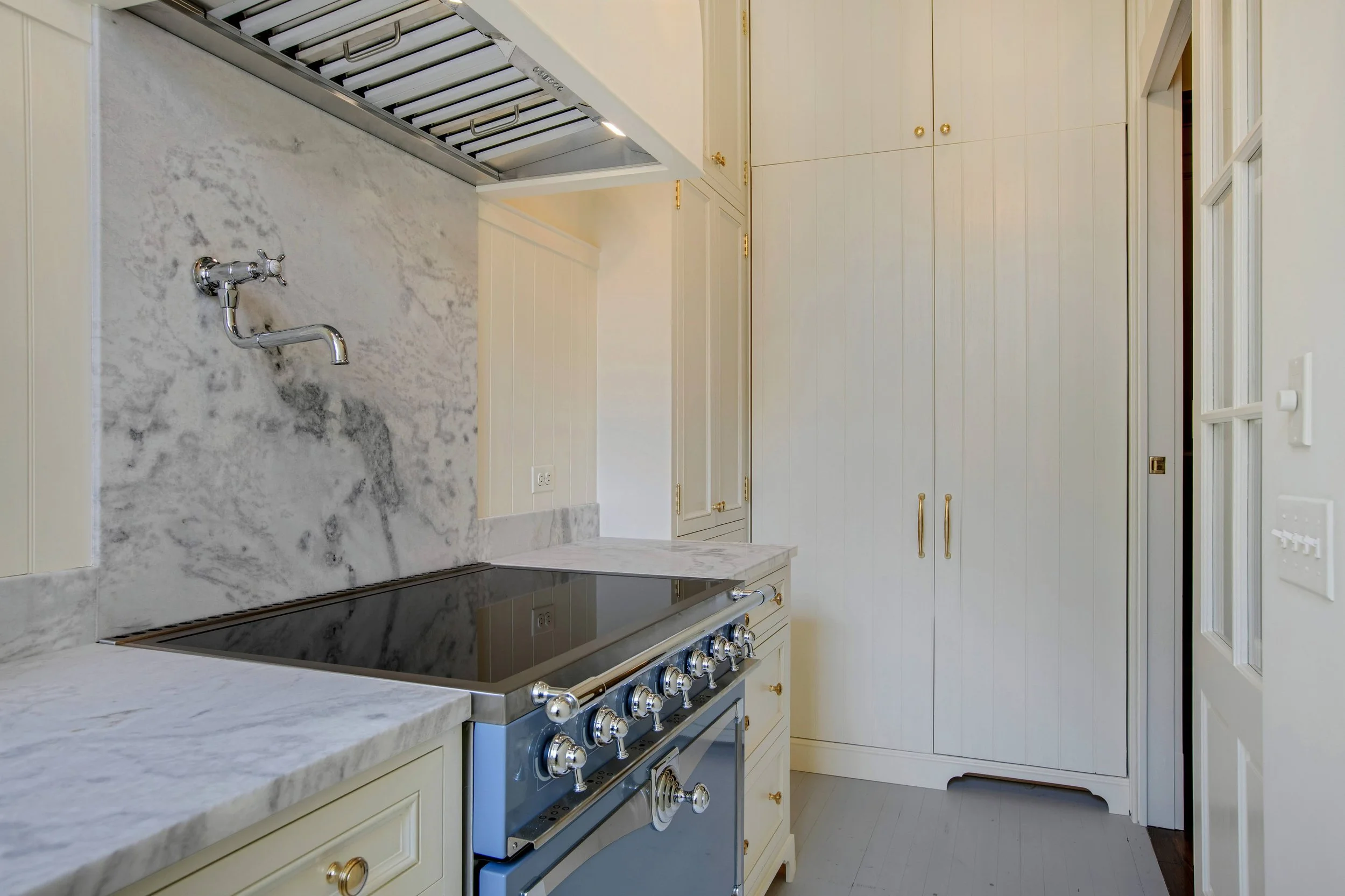 Kitchen with marble countertops, a stove with control knobs, cream-colored cabinets with gold handles, and a wall-mounted faucet.