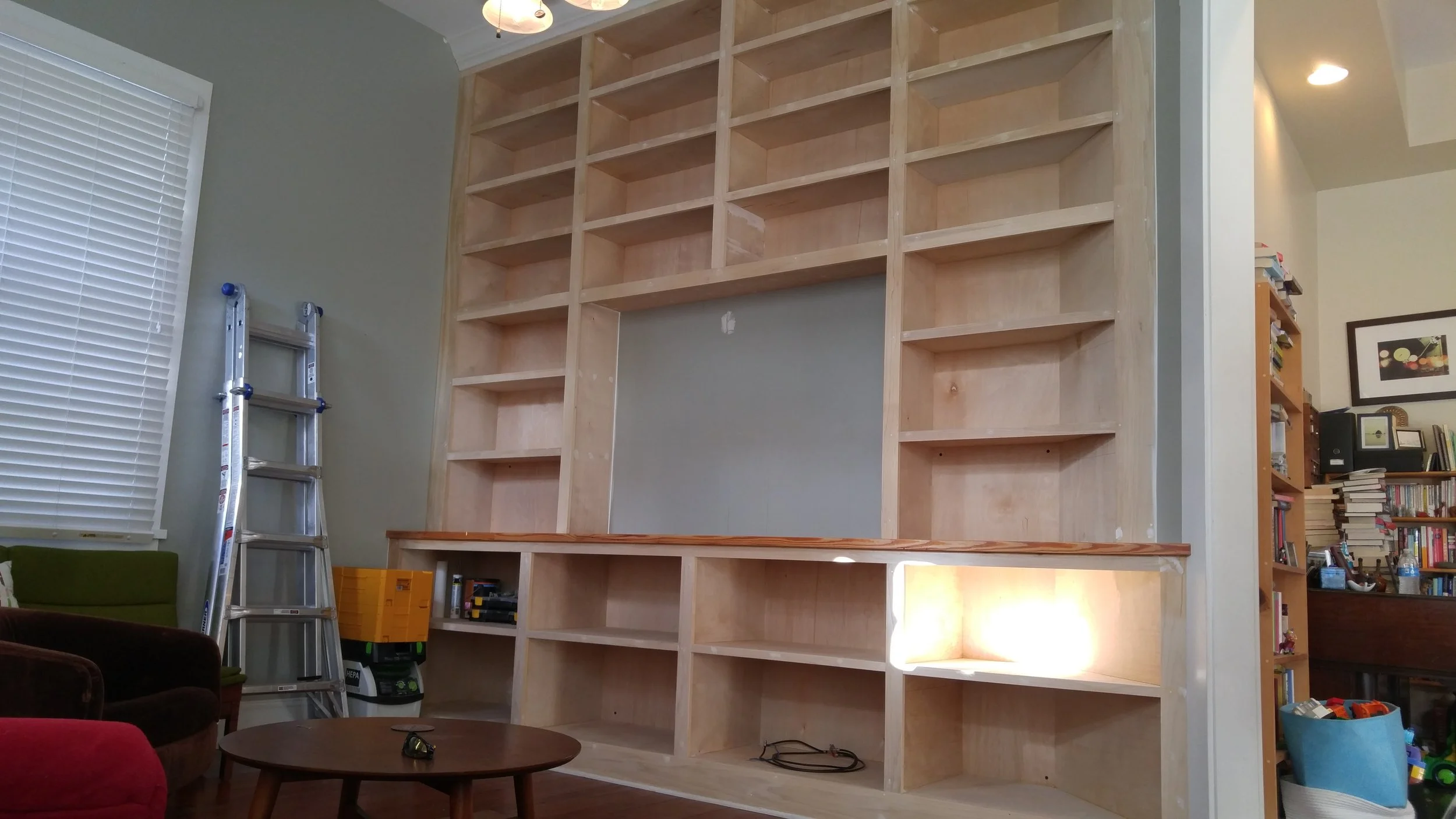 Room with an unfinished wooden built-in  entertainment center under construction, with shelves and compartments, next to a window with blinds, and a ladder leaning against the wall. .