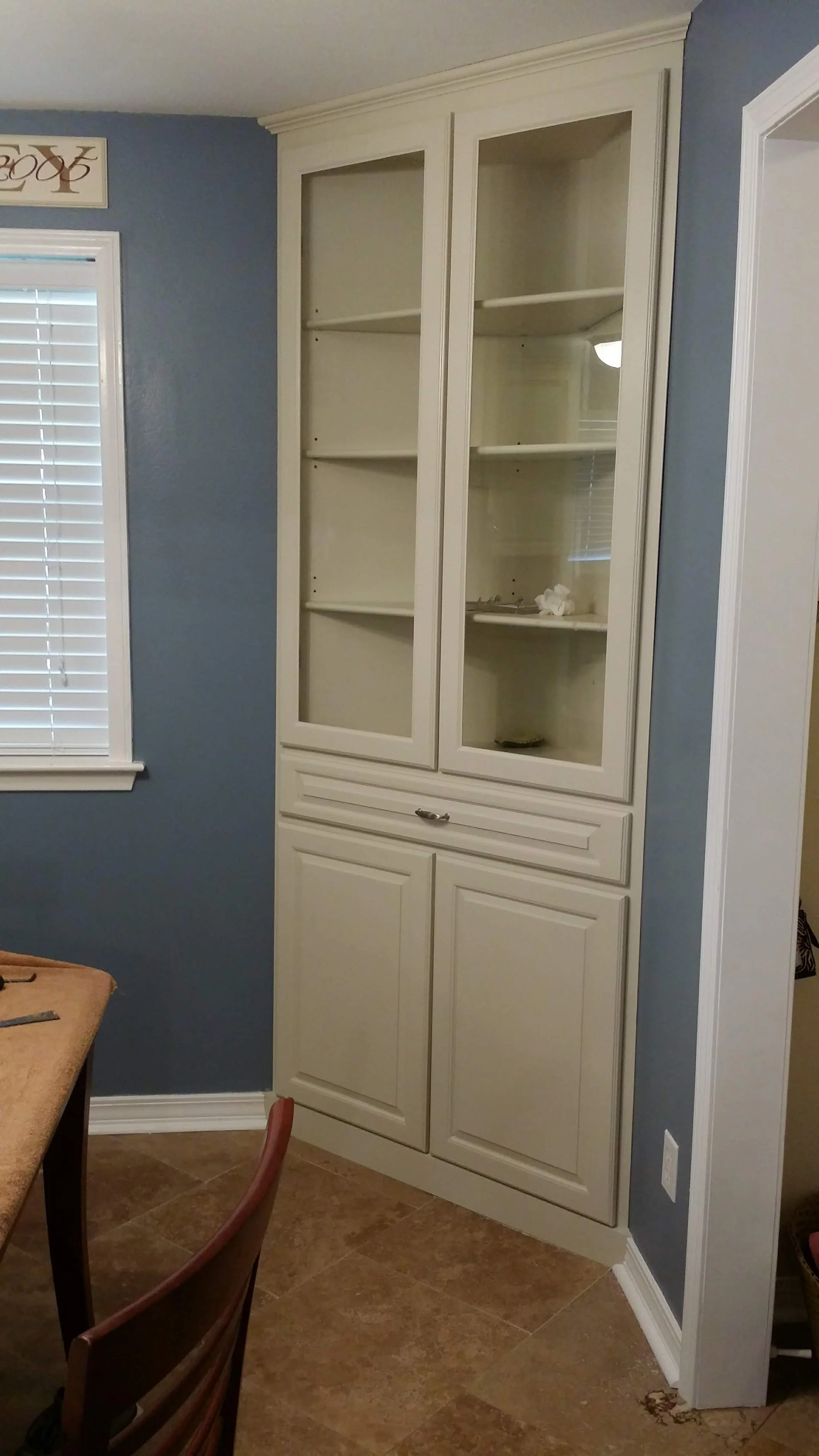 White built-in china cabinet with glass doors and closed cabinets below in a room with blue walls and tile flooring, next to a window with blinds.