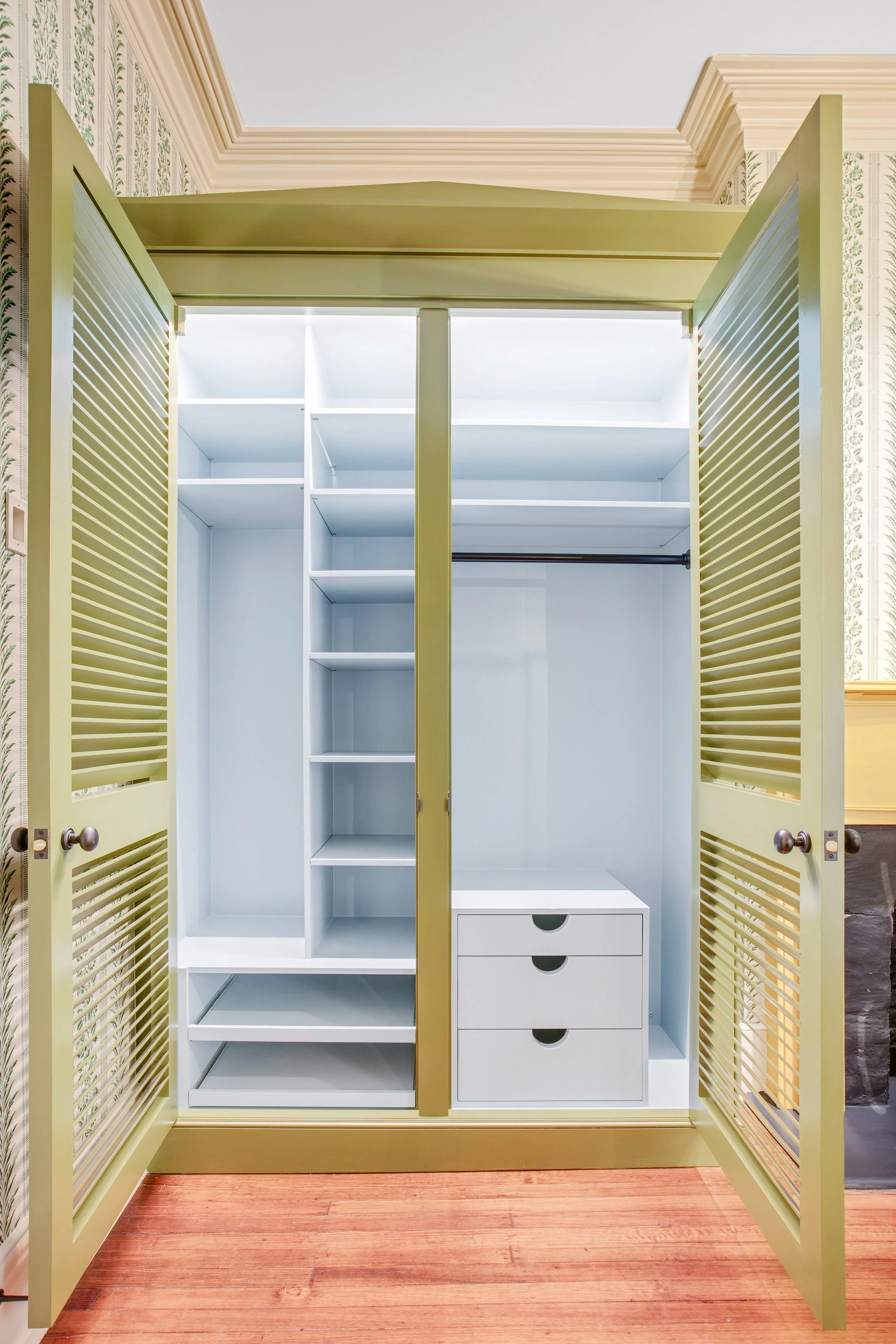Custom built-in armoire with yellow louvered doors, white interior, shelves, drawers, and hanging rod, on hardwood floor.