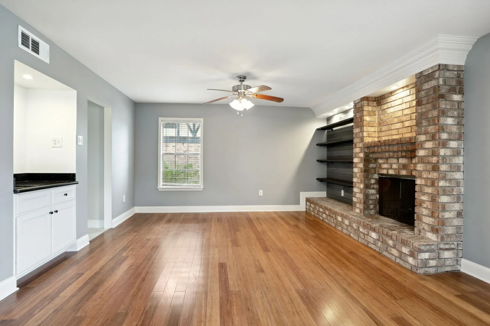 Empty living room with hardwood floors, gray walls, a ceiling fan, a brick fireplace with black shelves, a window, and a small built-in white cabinet.
