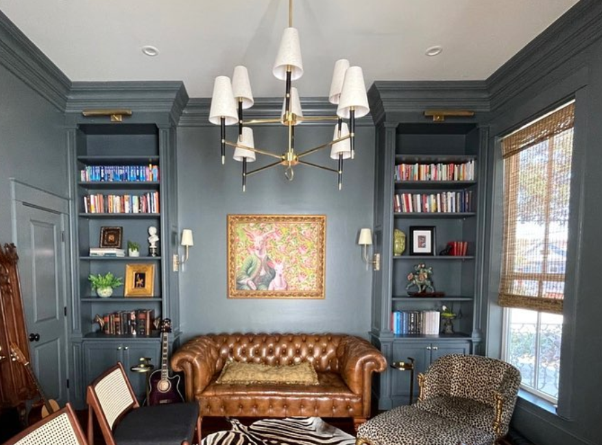 Living room with blue walls, built-in bookshelves filled with books and decorative items, a brown tufted leather sofa, a patterned armchair, a zebra-print rug, a window with bamboo shades, and a modern chandelier.