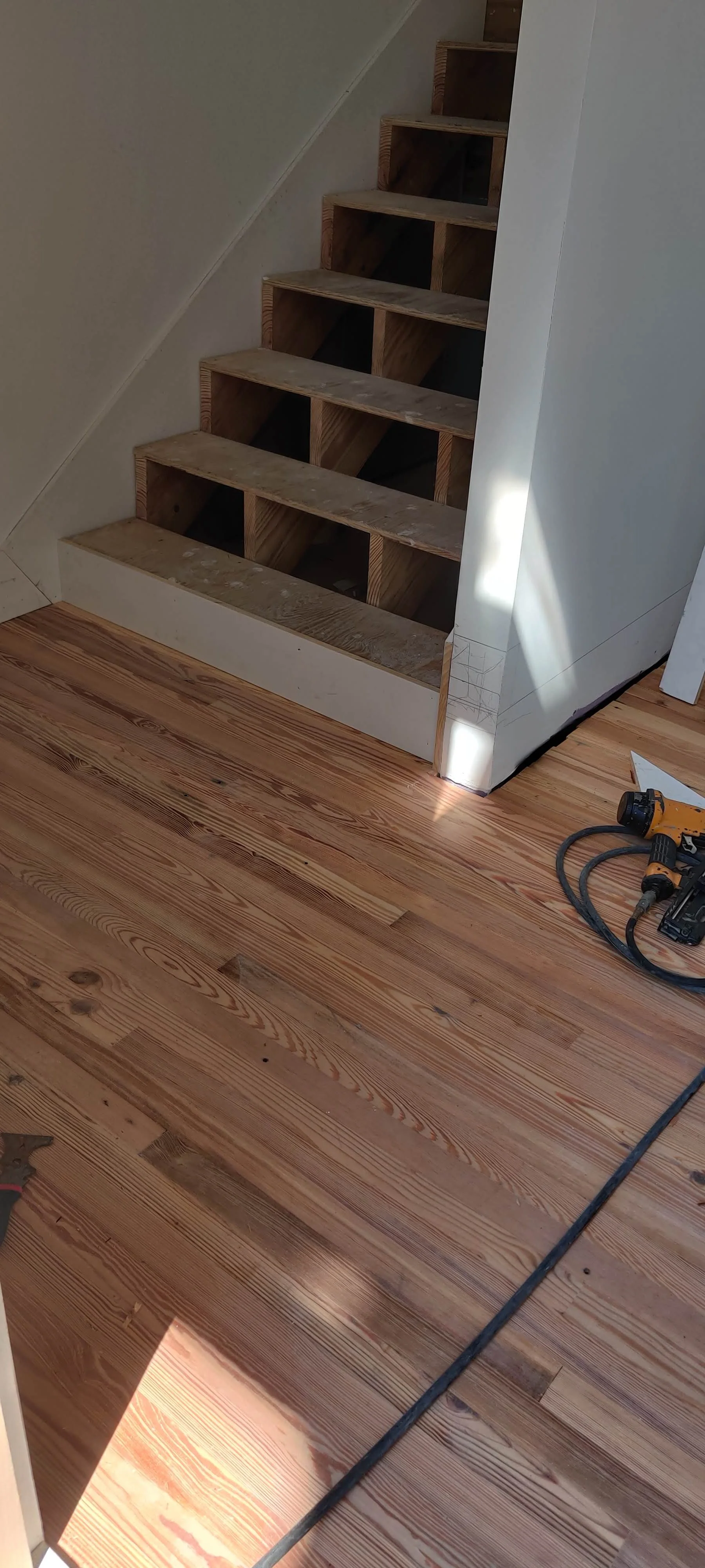 A staircase with unfinished wooden steps under construction, next to a room with hardwood flooring, construction tools on the floor, and white walls.