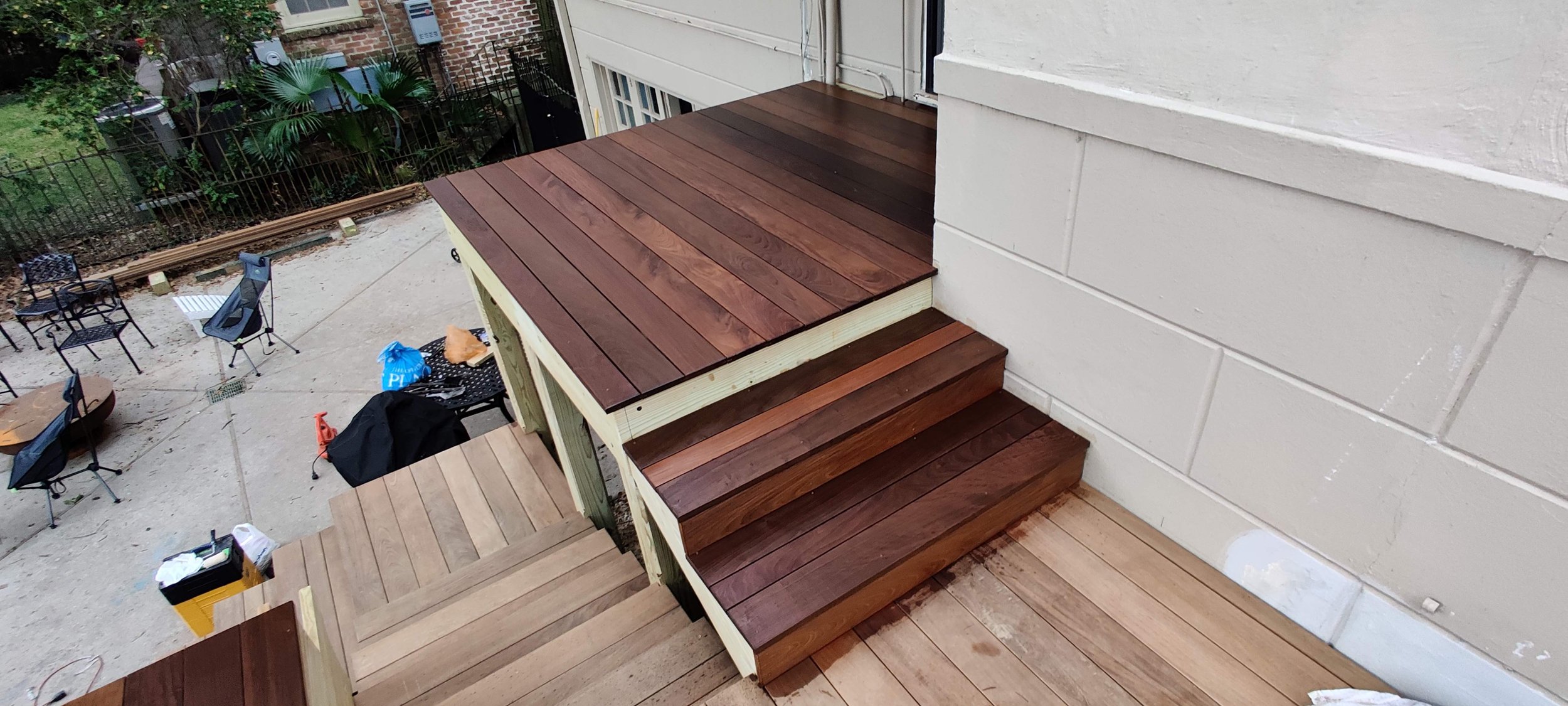 Photo of newly installed wooden stairs and deck outside a house, with outdoor furniture and tools visible on the ground.