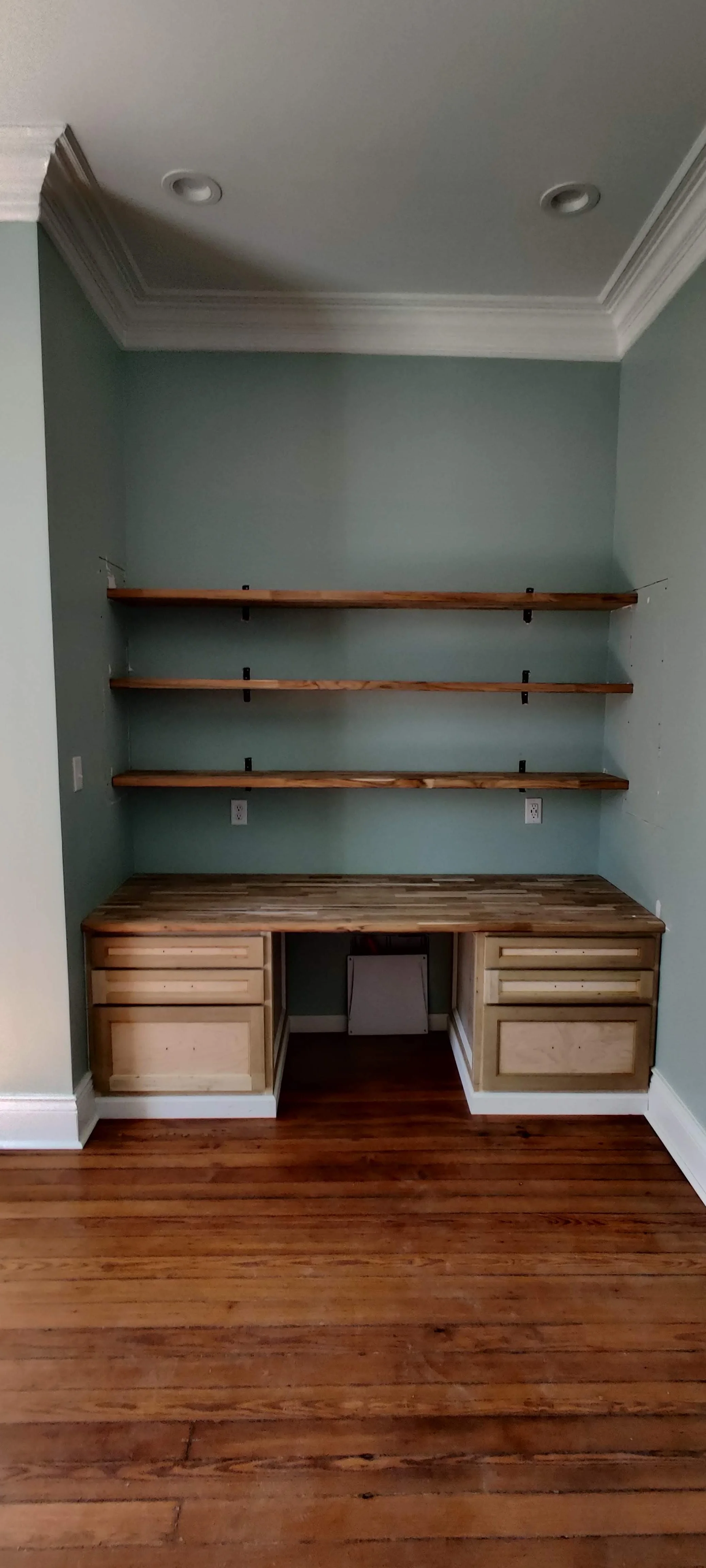 Empty built-in desk with three wooden shelves above and two drawers on each side, situated against a light blue wall in a room with hardwood floors.