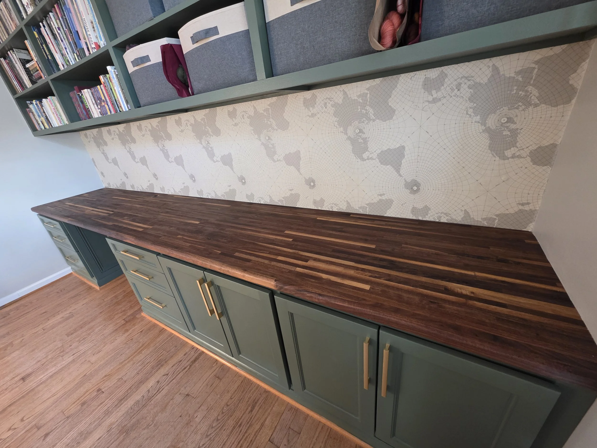 A built-in home office space with a green cabinet and a dark wood countertop, a map-themed backsplash, and gray shelves containing books and storage bins with yarn.