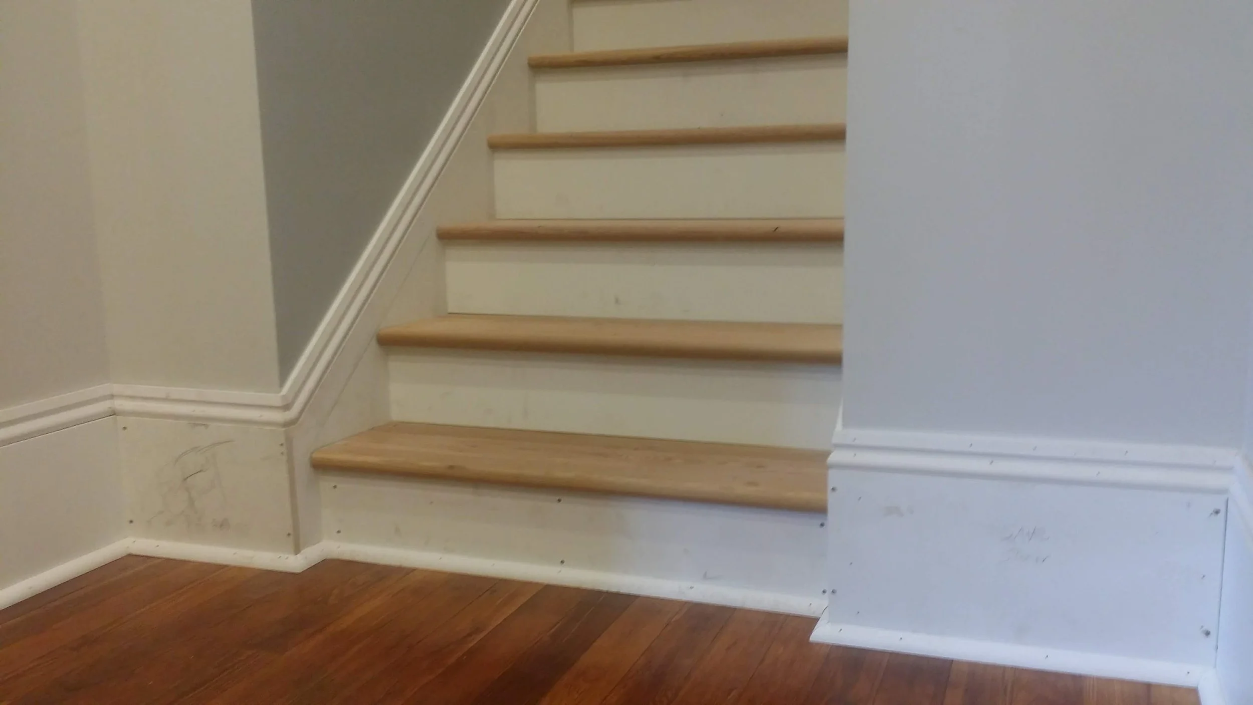 Interior view of a staircase with wooden steps and white risers, beige walls, and hardwood flooring.