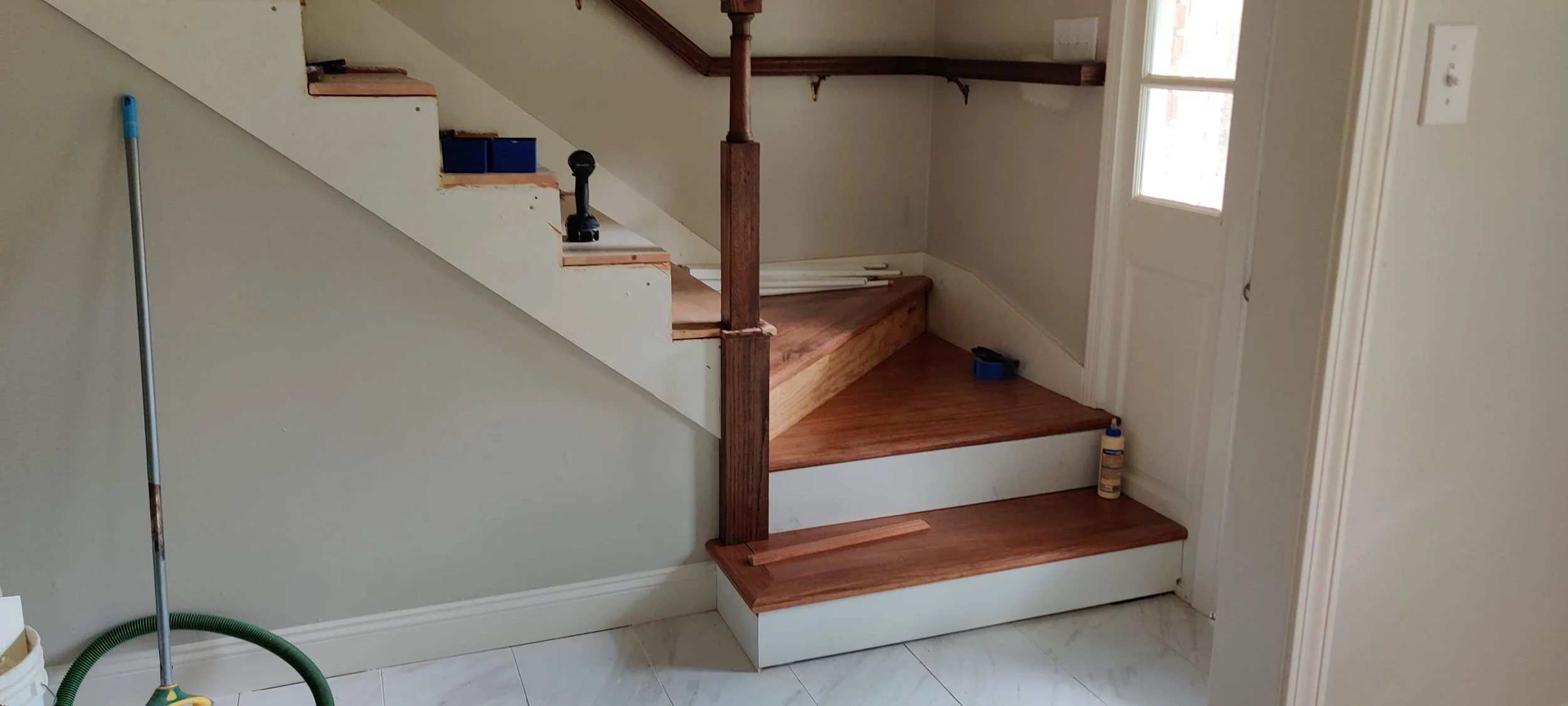 Interior of a house under renovation, showing a staircase with wooden steps, a white wall, and tools like a drill and a bottle of wood glue on the stairs.