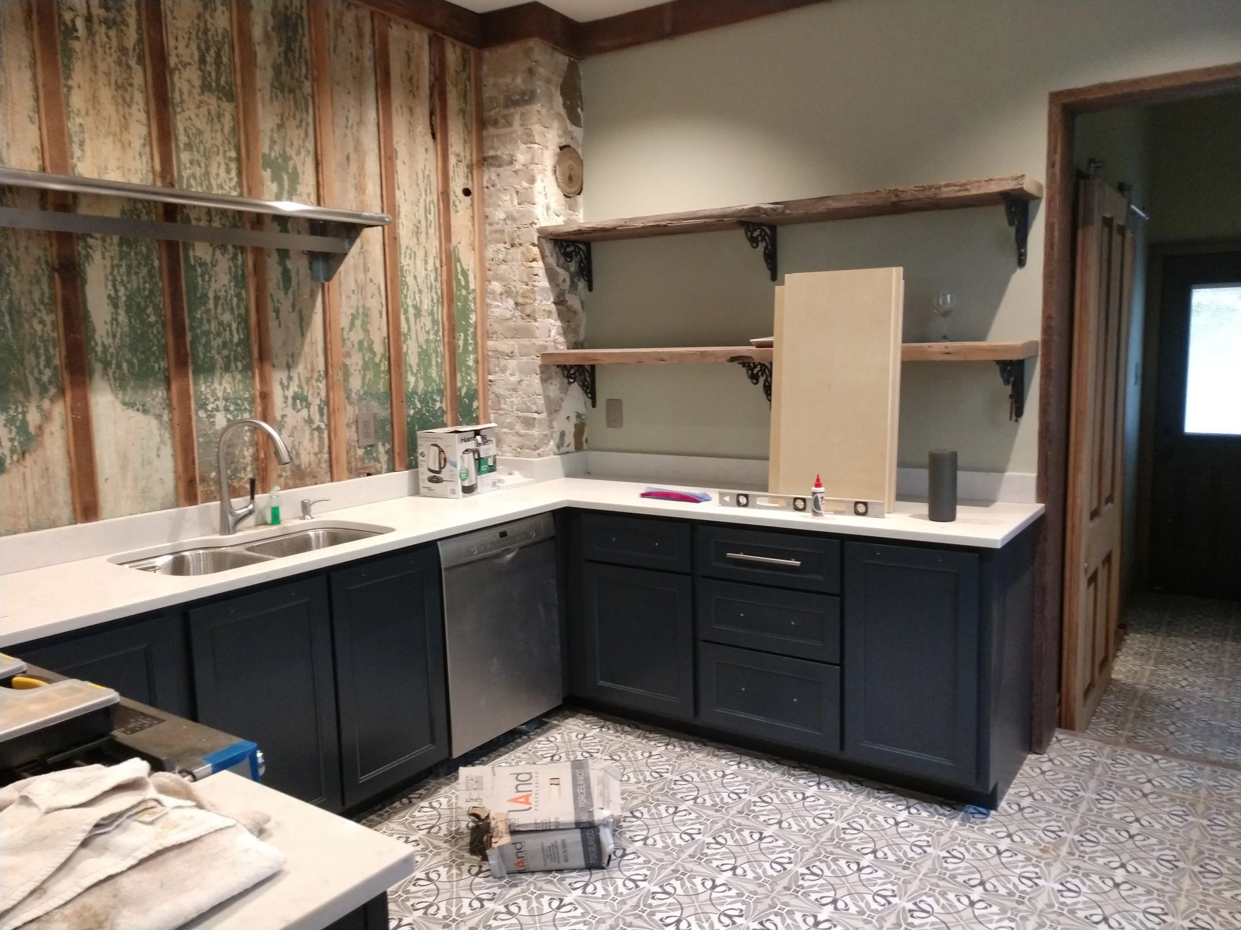 Kitchen with black cabinets, white countertop, and an unfinished wall with exposed wood and bricks. Shelves are installed on the wall, and various tools and materials are present, indicating ongoing renovation.