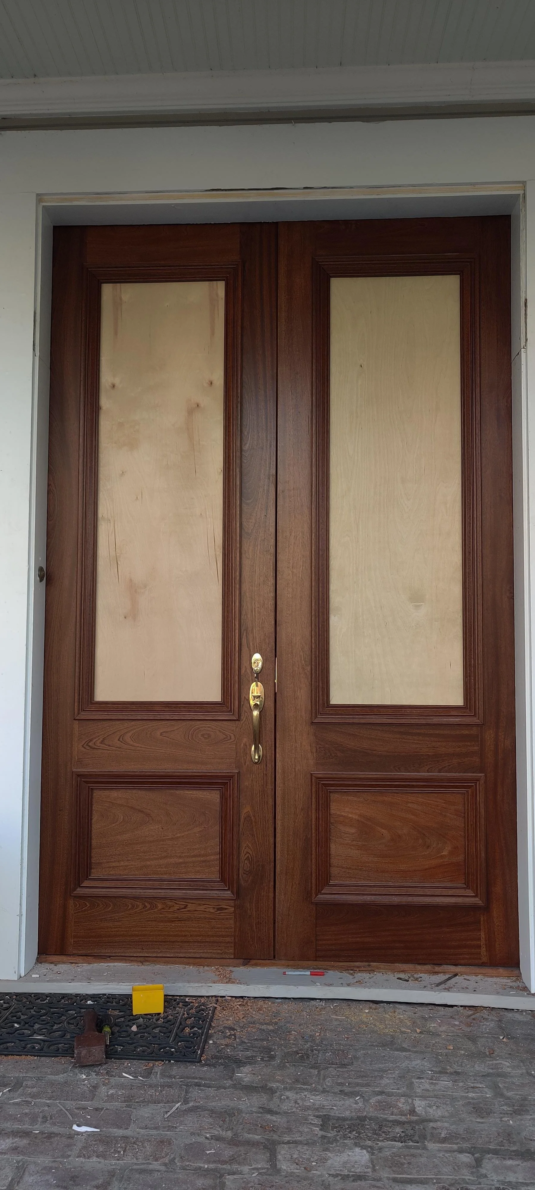 Wooden double front door with ornate molding, brass handles, and a lock, set in a white frame under a white ceiling, with construction materials on the brick porch.