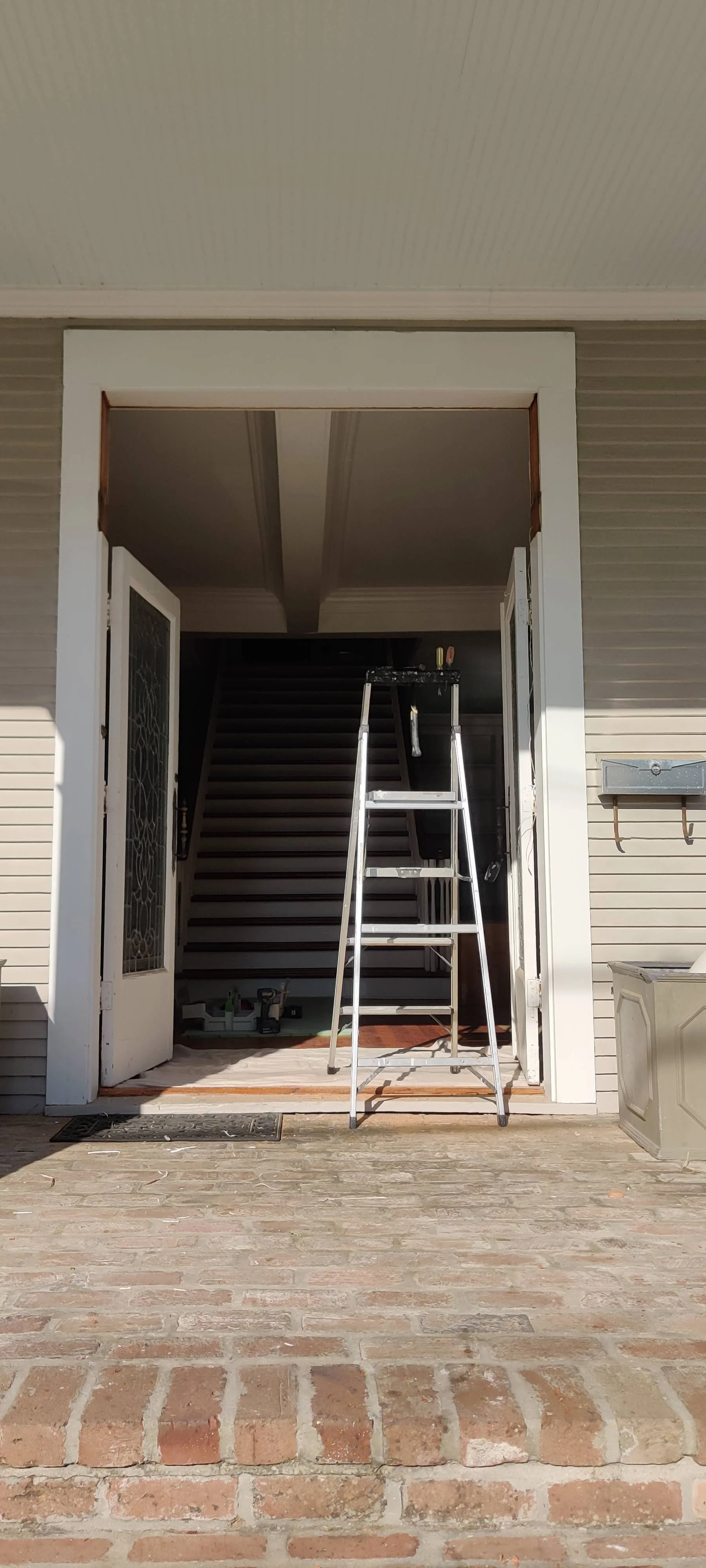 House entryway with open door, ladder, staircase inside, brick porch, mailbox, and toolbox inside.