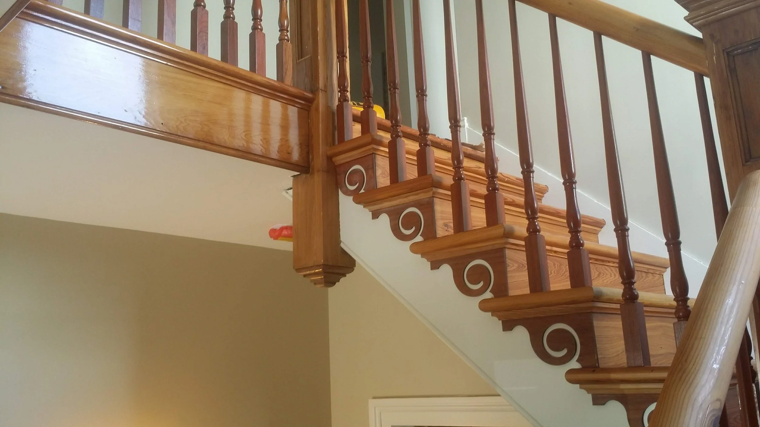A wooden staircase with ornate scroll details on the side, wooden spindles, and a polished handrail, seen from below inside a house.