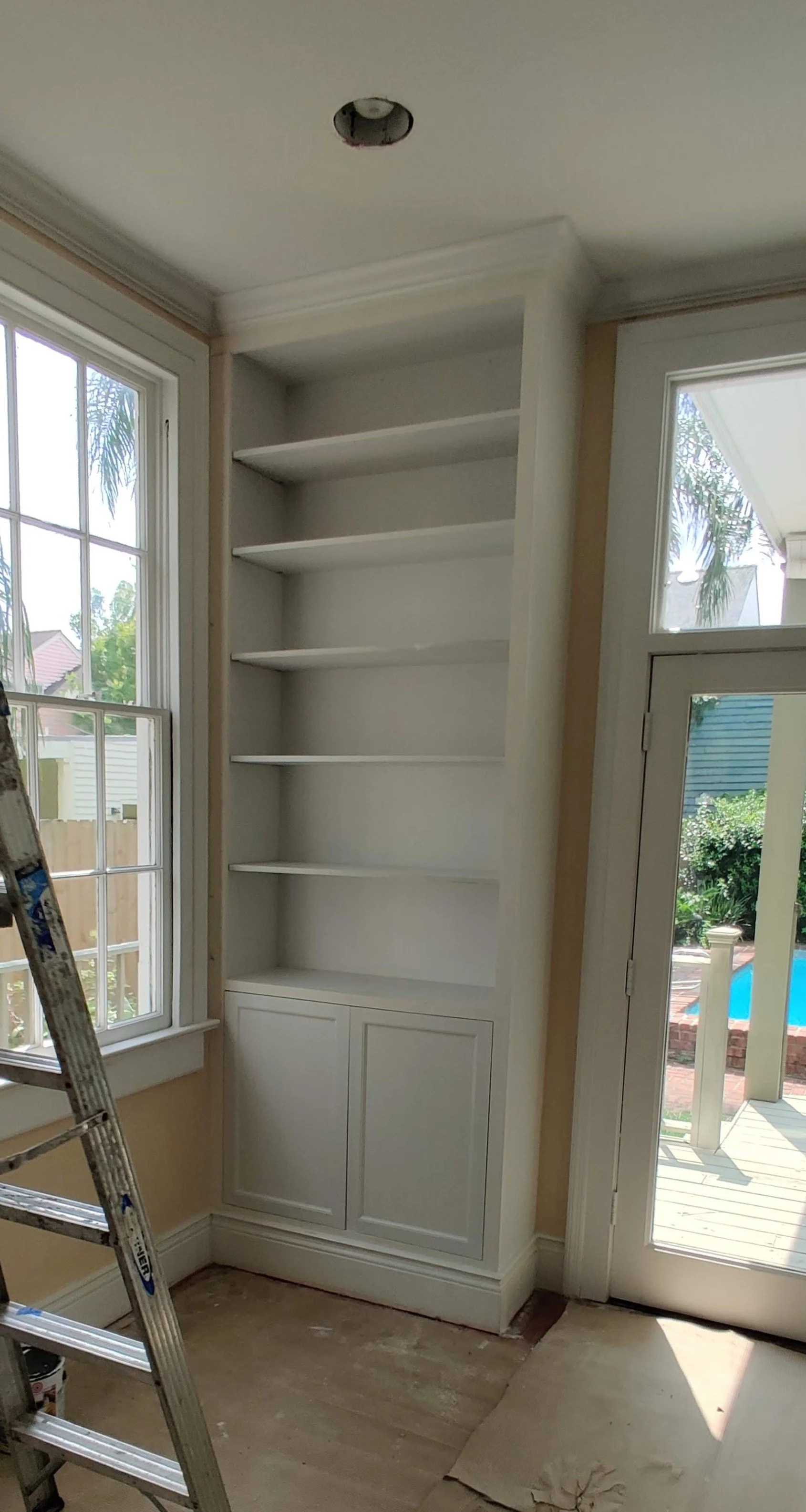 An empty white built-in bookshelf with five shelves and a closed cabinet at the bottom, located next to a window and a glass door leading outside, in a room under renovation.