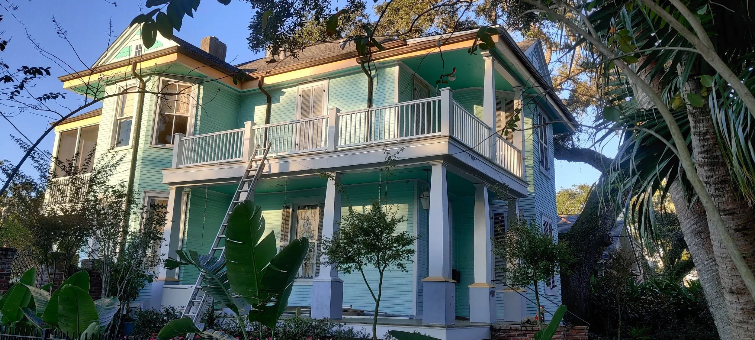 A light blue, multi-story house with a wrap-around balcony, surrounded by trees and tropical plants, under a clear blue sky.