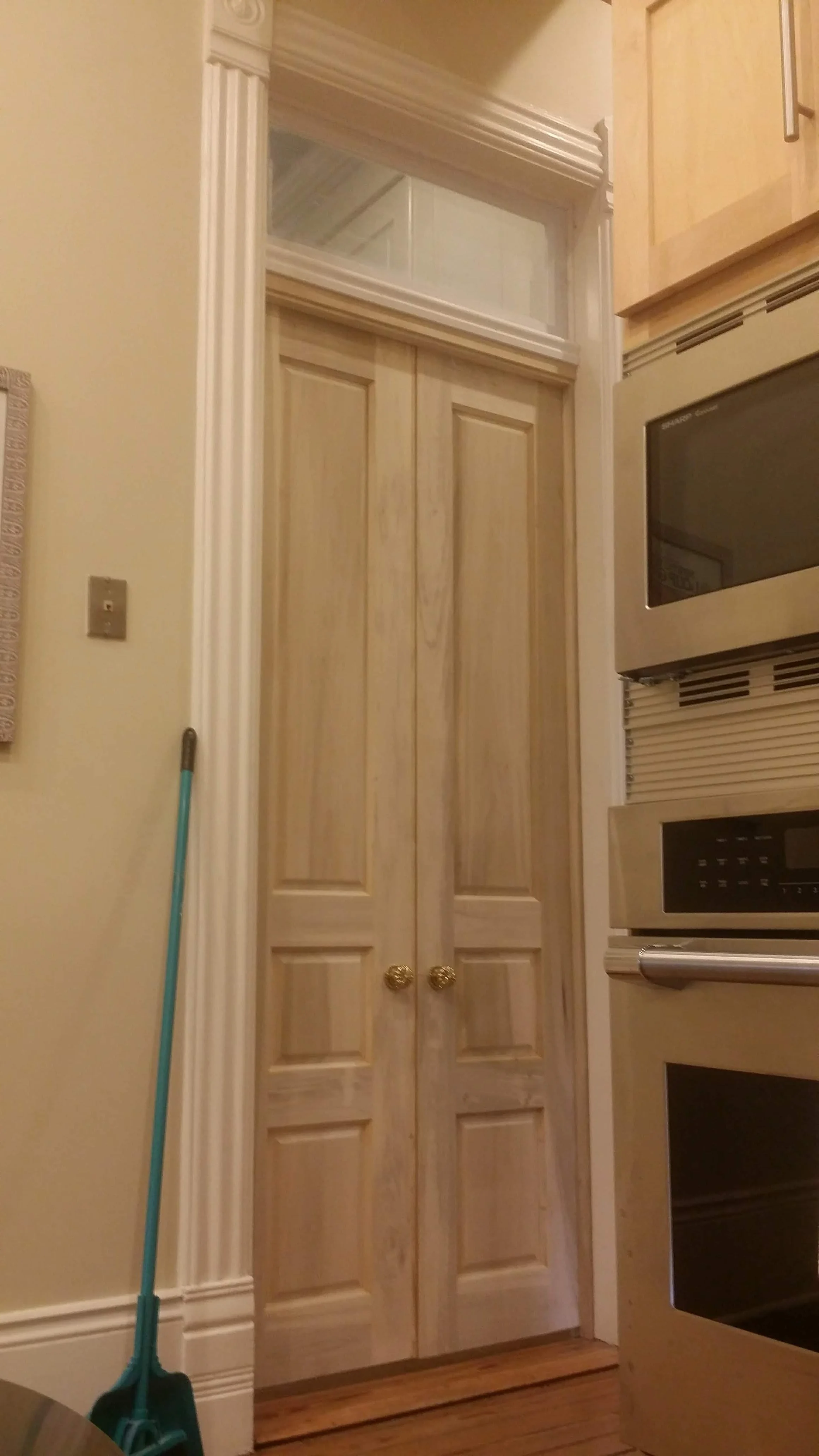 Kitchen with beige wooden double doors, a broom leaned against the wall, and stainless steel microwave and oven.