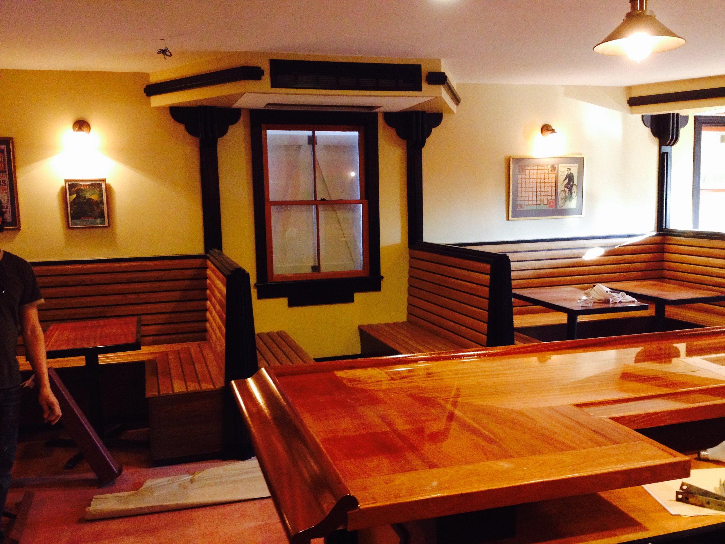 Empty restaurant with wooden booths and tables, a window with black trim, framed artwork on the walls, and warm lighting fixtures.