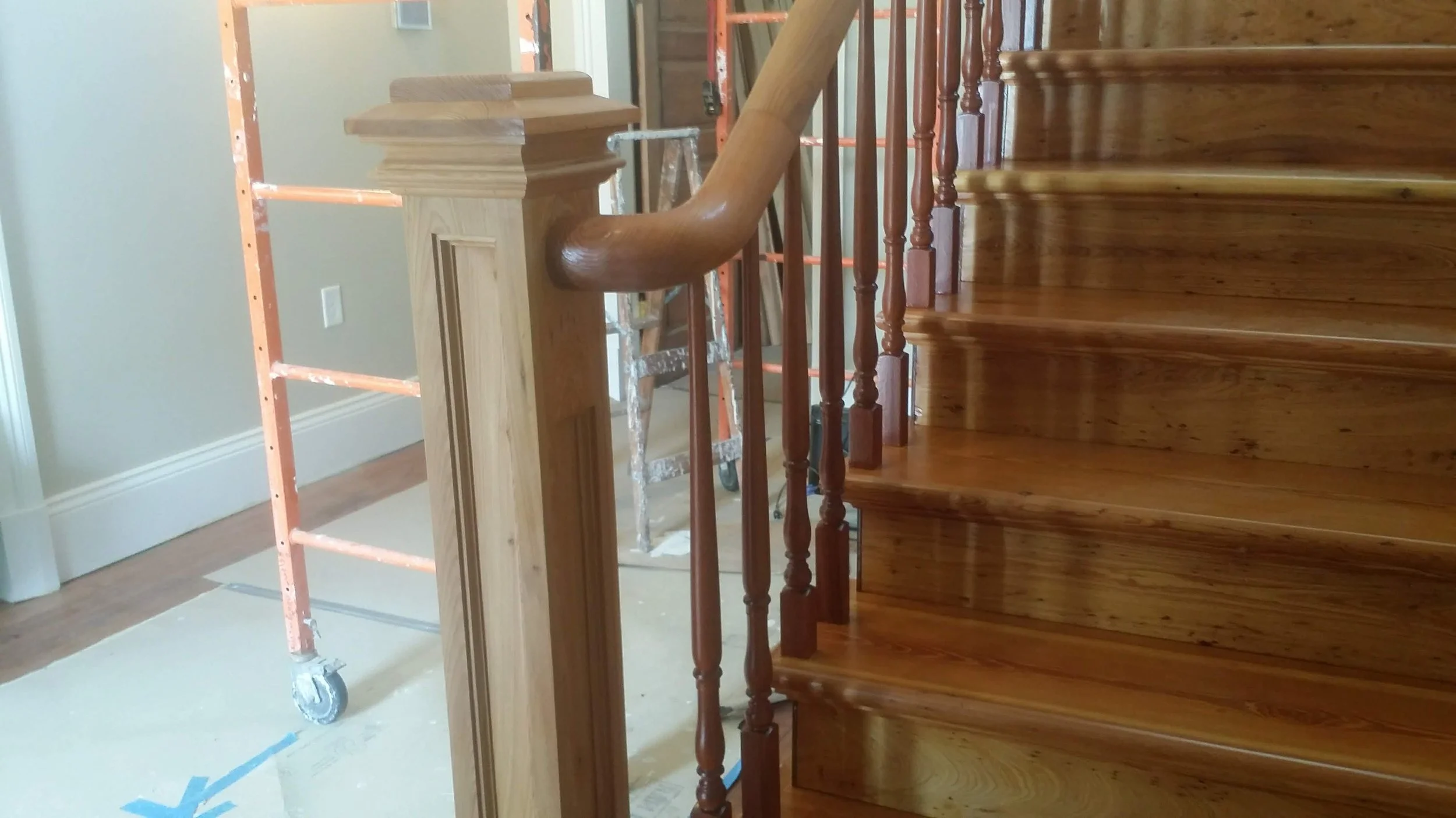 Wooden staircase with a polished handrail and spindles, under construction with a scaffolding and tools visible in the background.